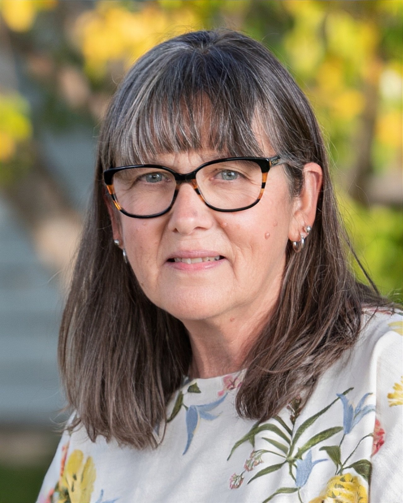 A smiling woman with gray hair and glasses wearing a black top and necklace, against a background of green foliage.