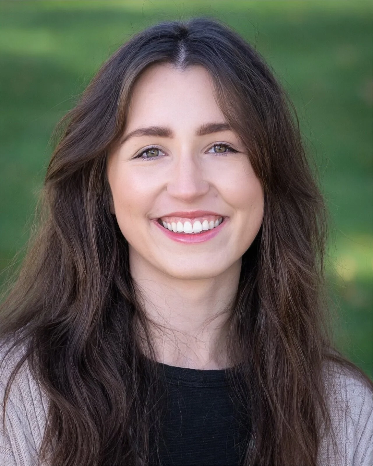 A young woman with wavy brown hair smiling at the camera in a pumpkin patch at sunset.