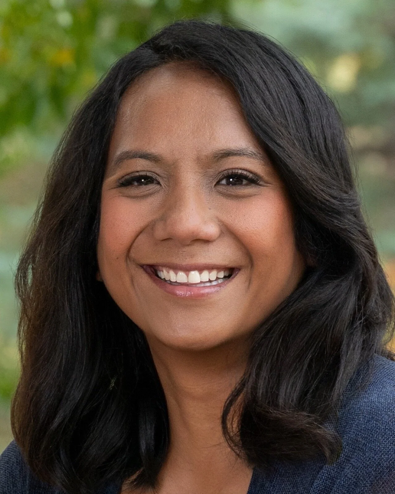 A woman with long dark hair, wearing a yellow top and a white earring, smiling in front of a wooden background.