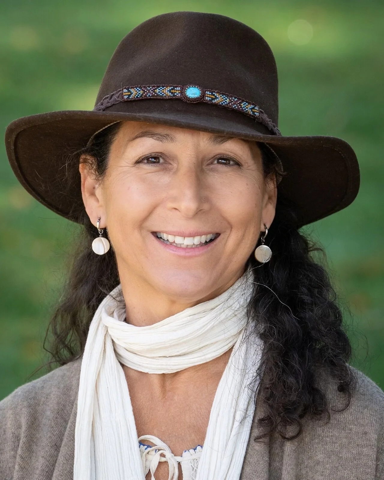 A smiling woman wearing a brown hat, teal scarf, and hoop earrings stands outdoors in a snowy mountain landscape.