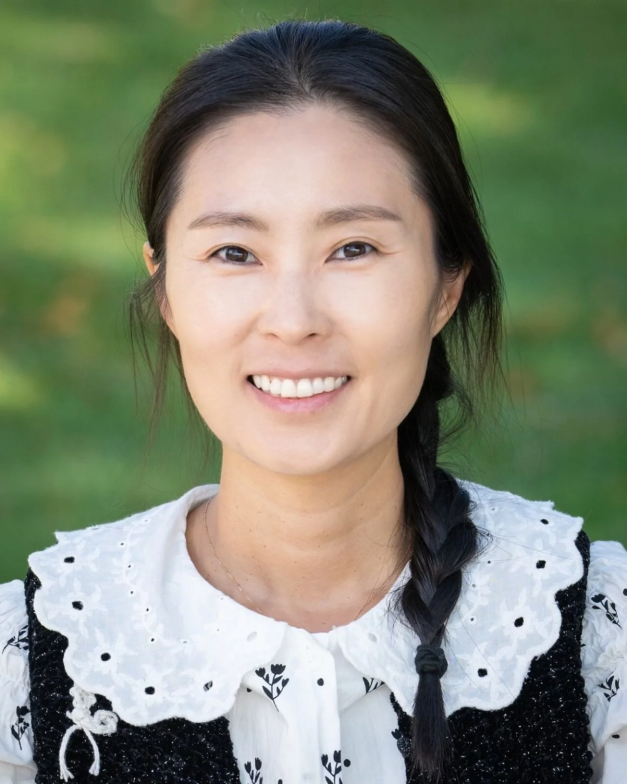A young woman with shoulder-length dark hair smiling, wearing a pale pink striped button-up shirt, against a brown and beige abstract background.