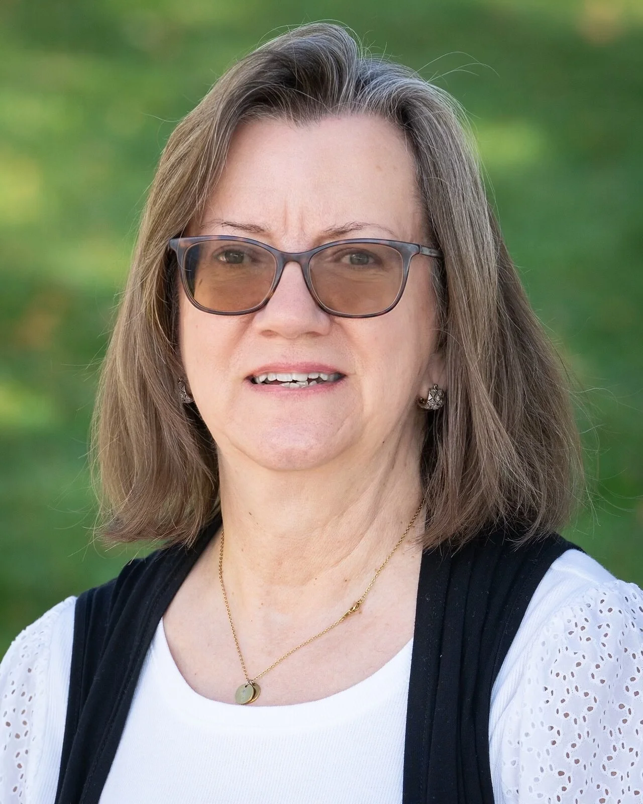 Woman with glasses smiling indoors, wearing a colorful scarf and blue top, in a room with wooden window frames and chairs in the background.