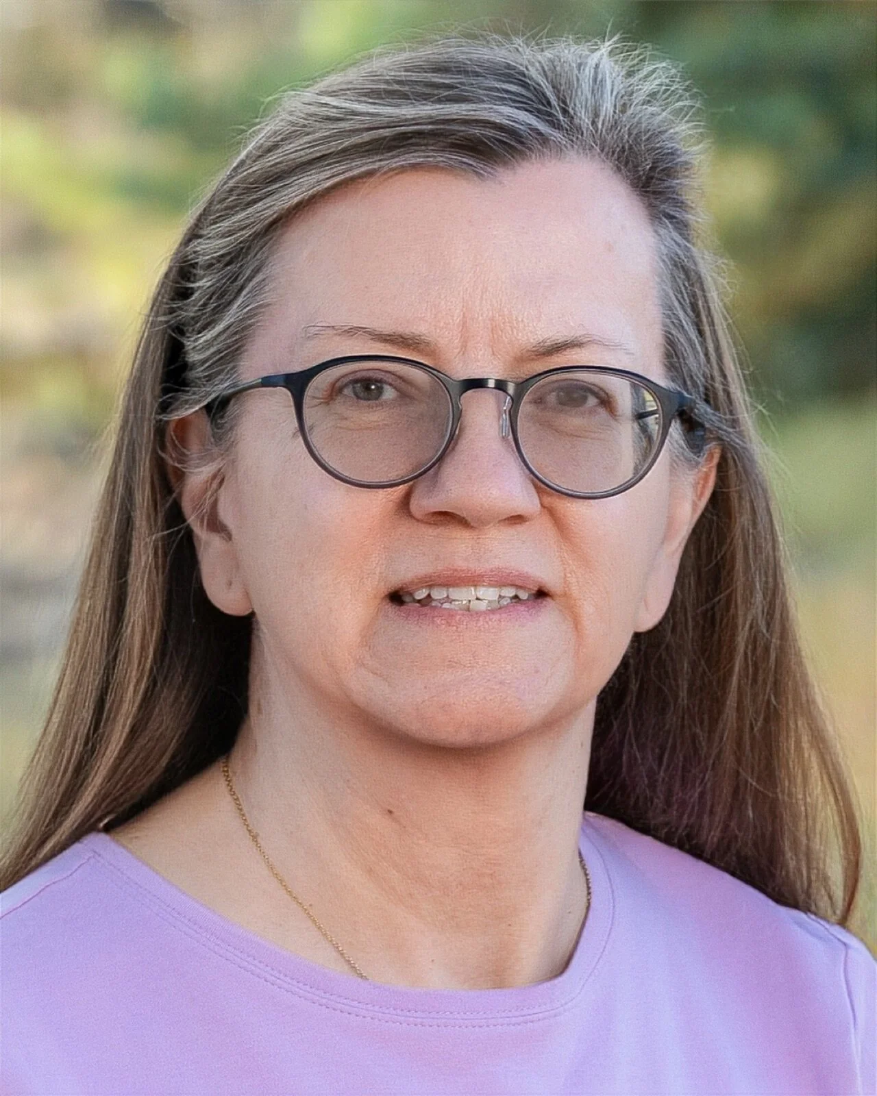 Woman with glasses smiling indoors, wearing a colorful scarf and blue top, in a room with wooden window frames and chairs in the background.