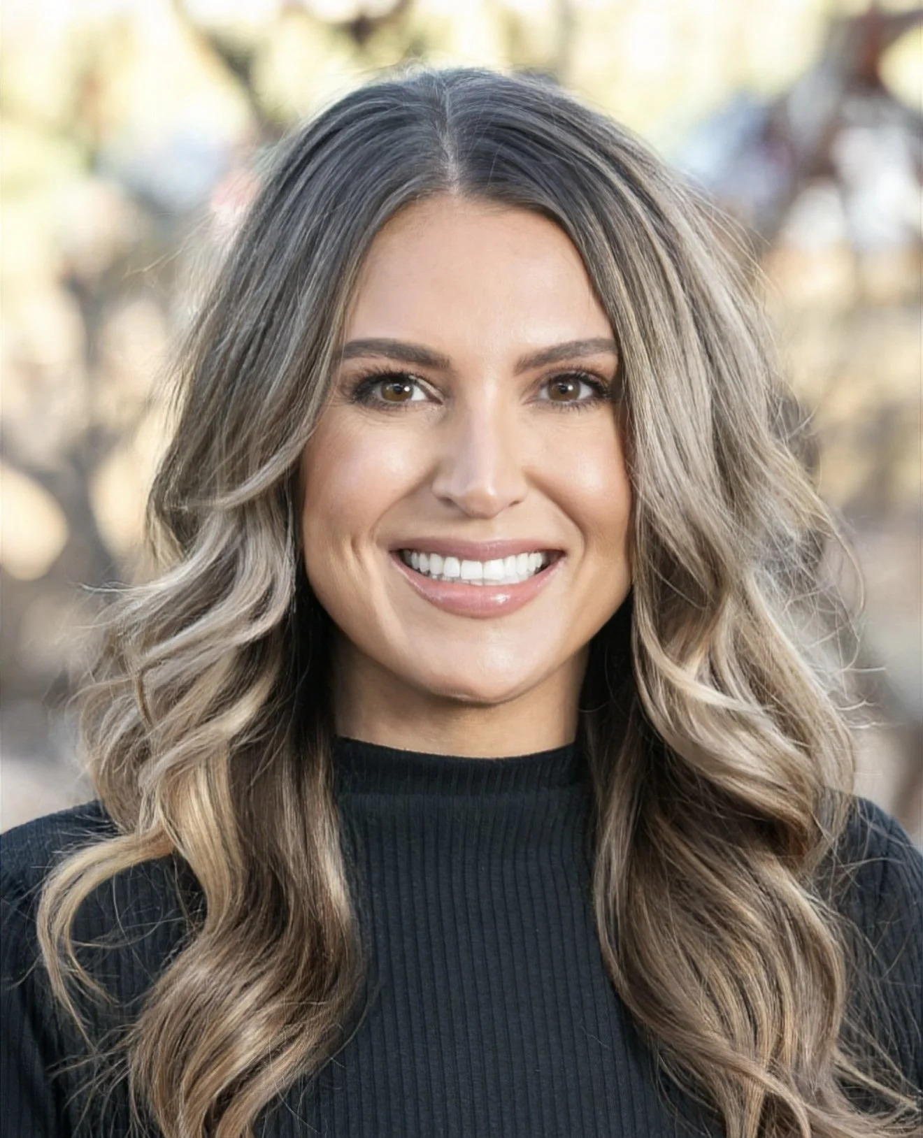 Smiling person with long blonde hair wearing a black top and earrings, against a white background.