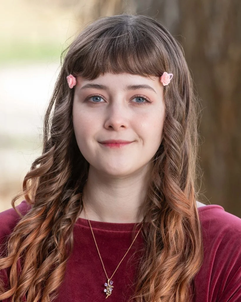 Smiling person outdoors in casual clothing, with a house and trees in the background.