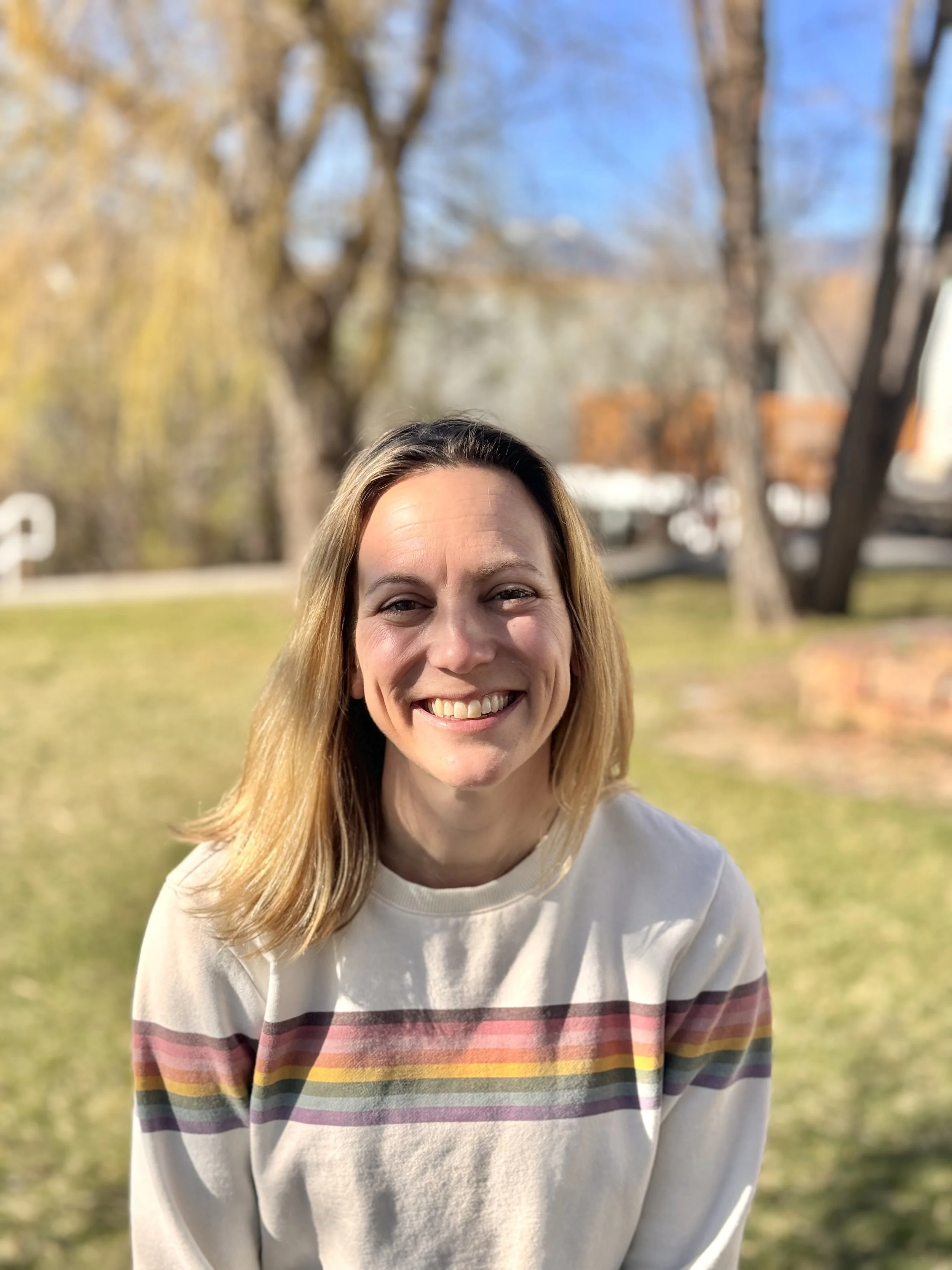 Smiling person in a white sweater with rainbow stripes, sitting outdoors with trees and grass in the background.