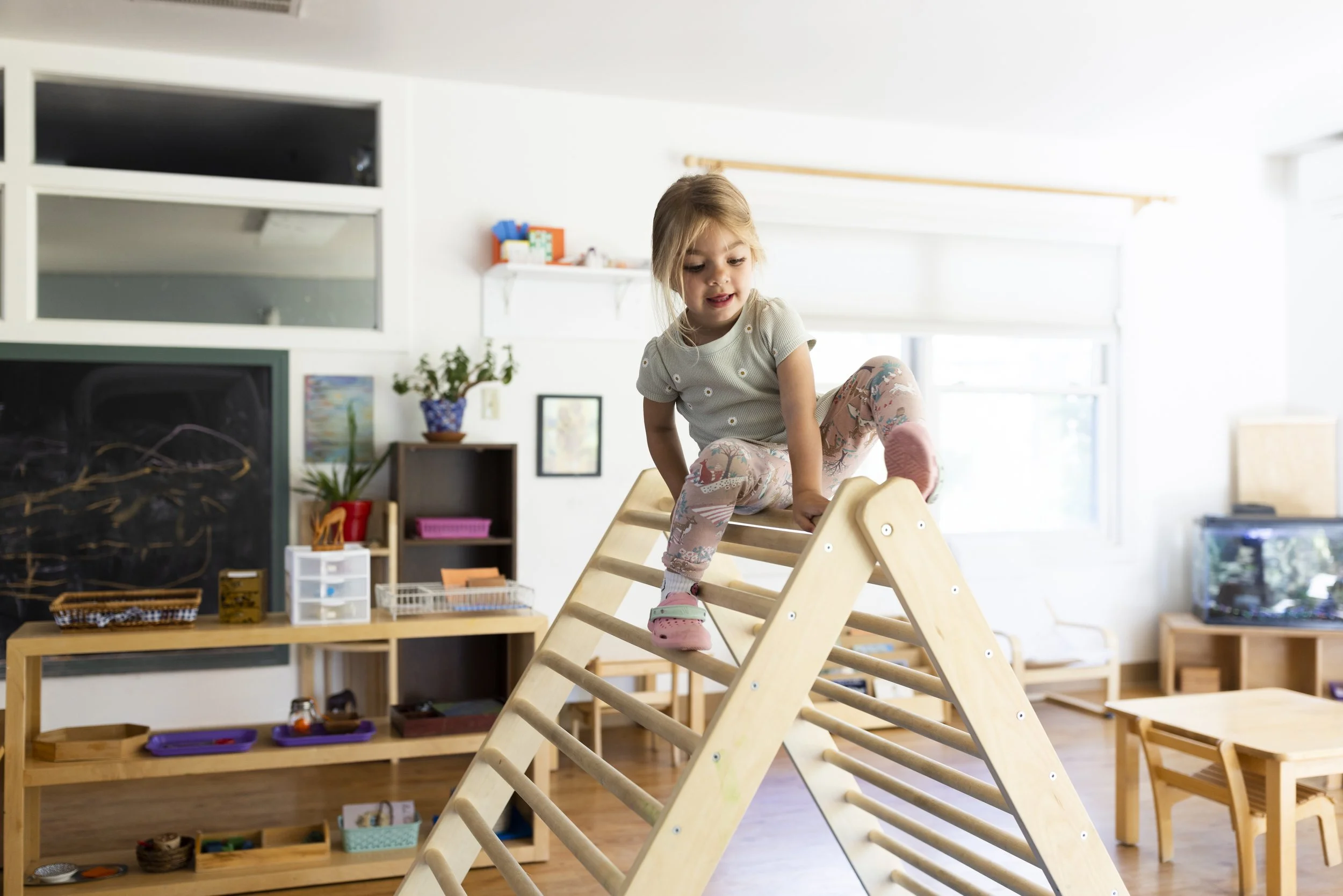 Young child practicing hand sewing with a needle and thread, using a red pincushion, in a classroom environment.