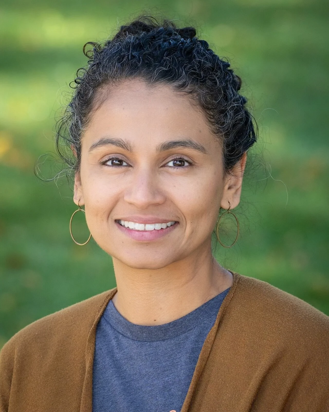 Smiling person with curly hair wearing a black top in front of greenery
