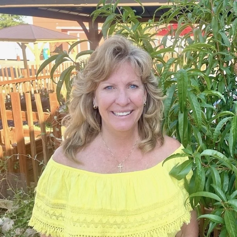 Woman in yellow top standing near green plants and wooden fence outdoors.