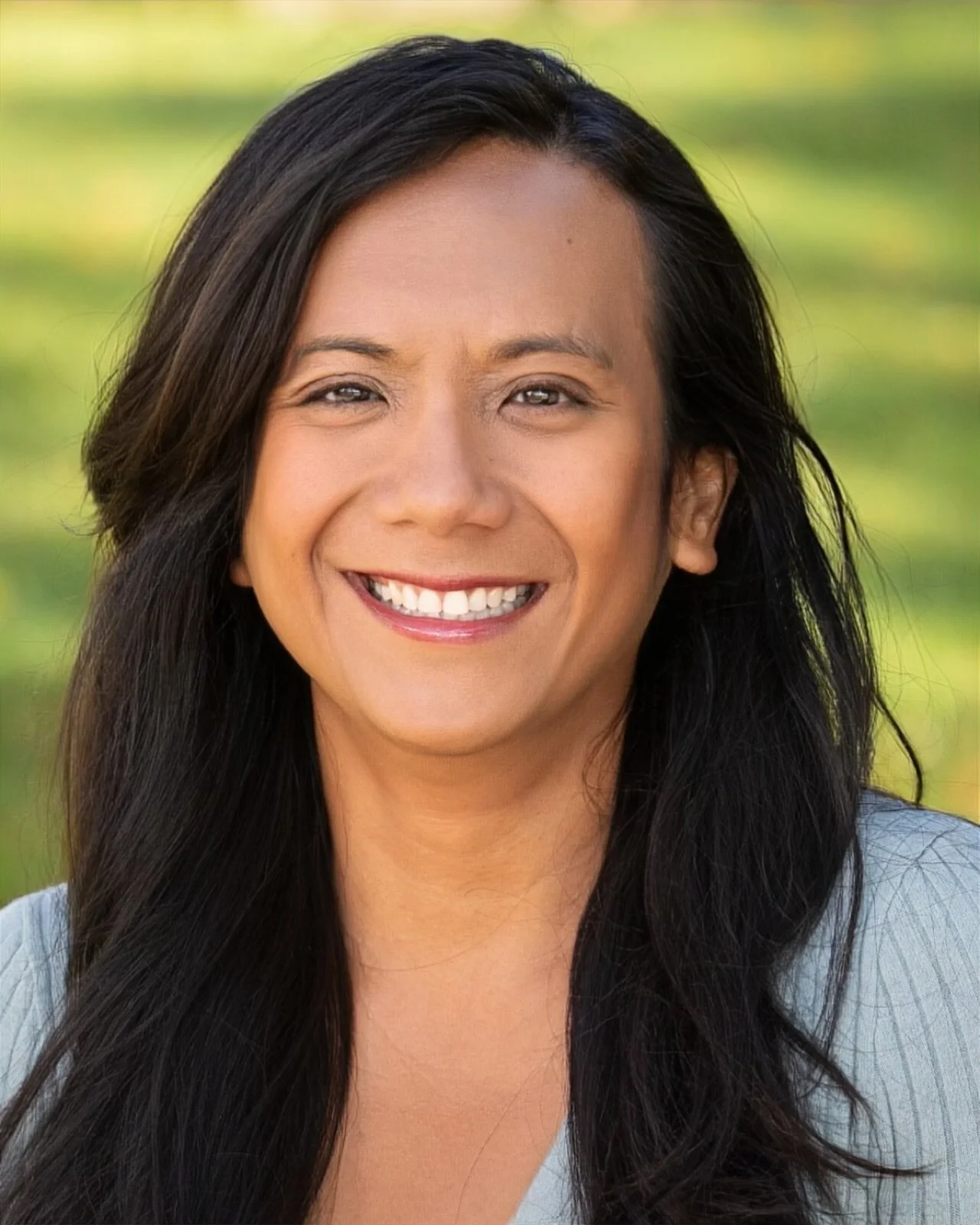 A woman with long dark hair, wearing a yellow top and a white earring, smiling in front of a wooden background.