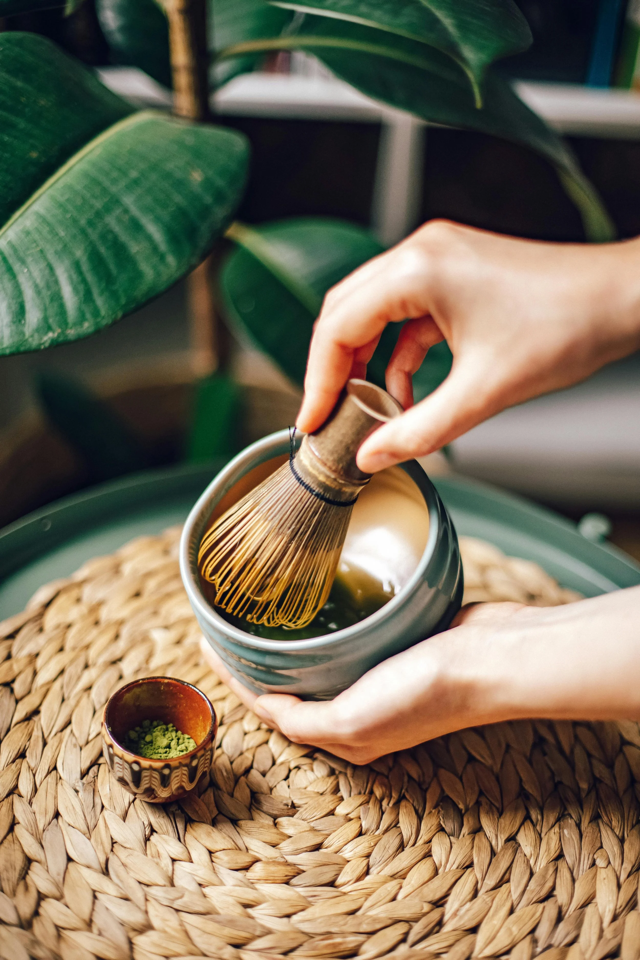A person uses a bamboo whisk to prepare matcha tea in a gray mug. There are green leaves background, a small decorative bowl with matcha powder, and a small cup with green powder on a woven placemat.