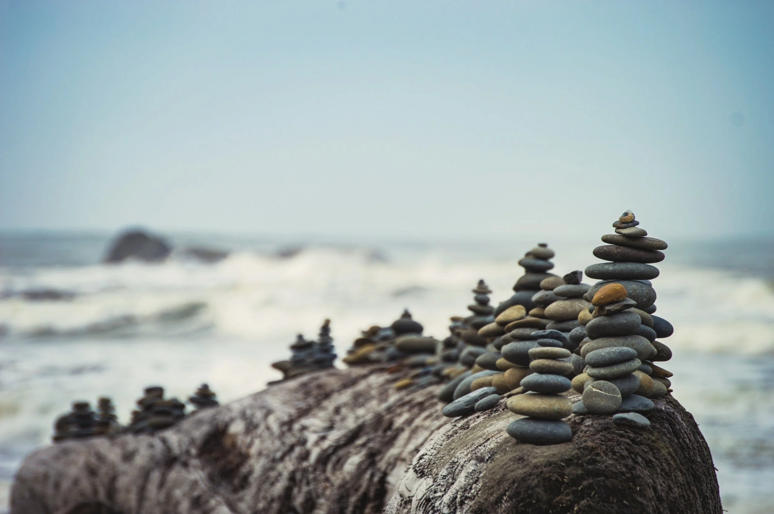 Stacks of smooth stones arranged along a log on a beach with the ocean in the background.