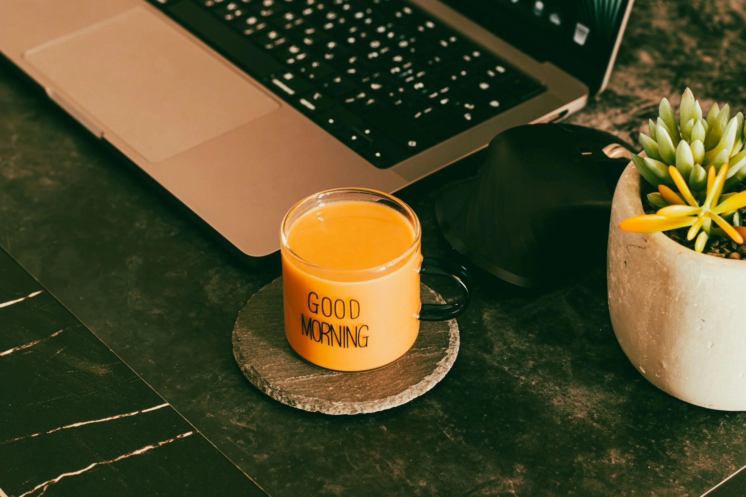 A laptop on a dark green marble table with a potted succulent plant and a glass mug of orange juice that says 'Good Morning'.