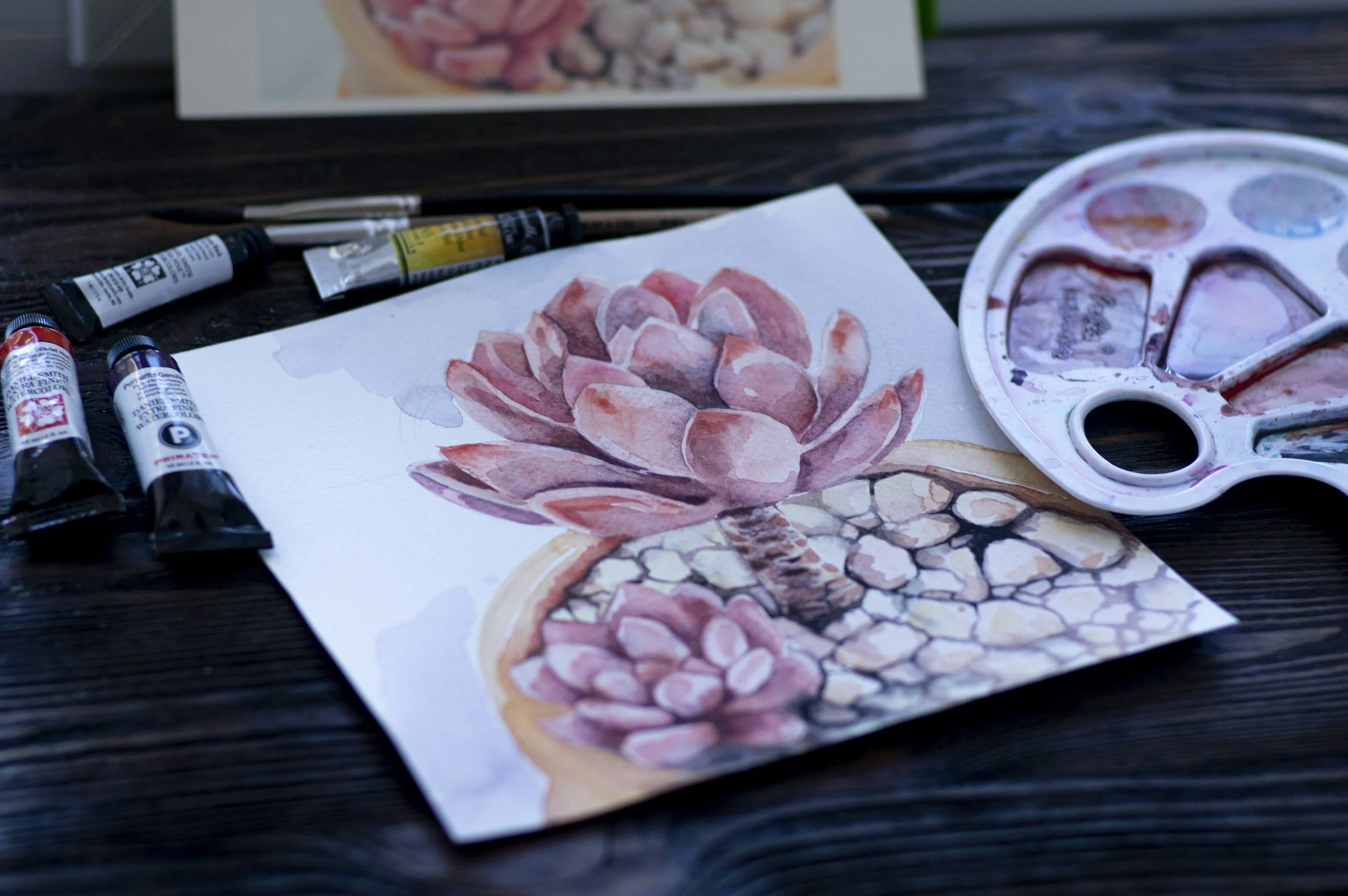 A watercolor painting of a pink succulent plant, with watercolor paints, pens, and a color palette on a dark wooden table.