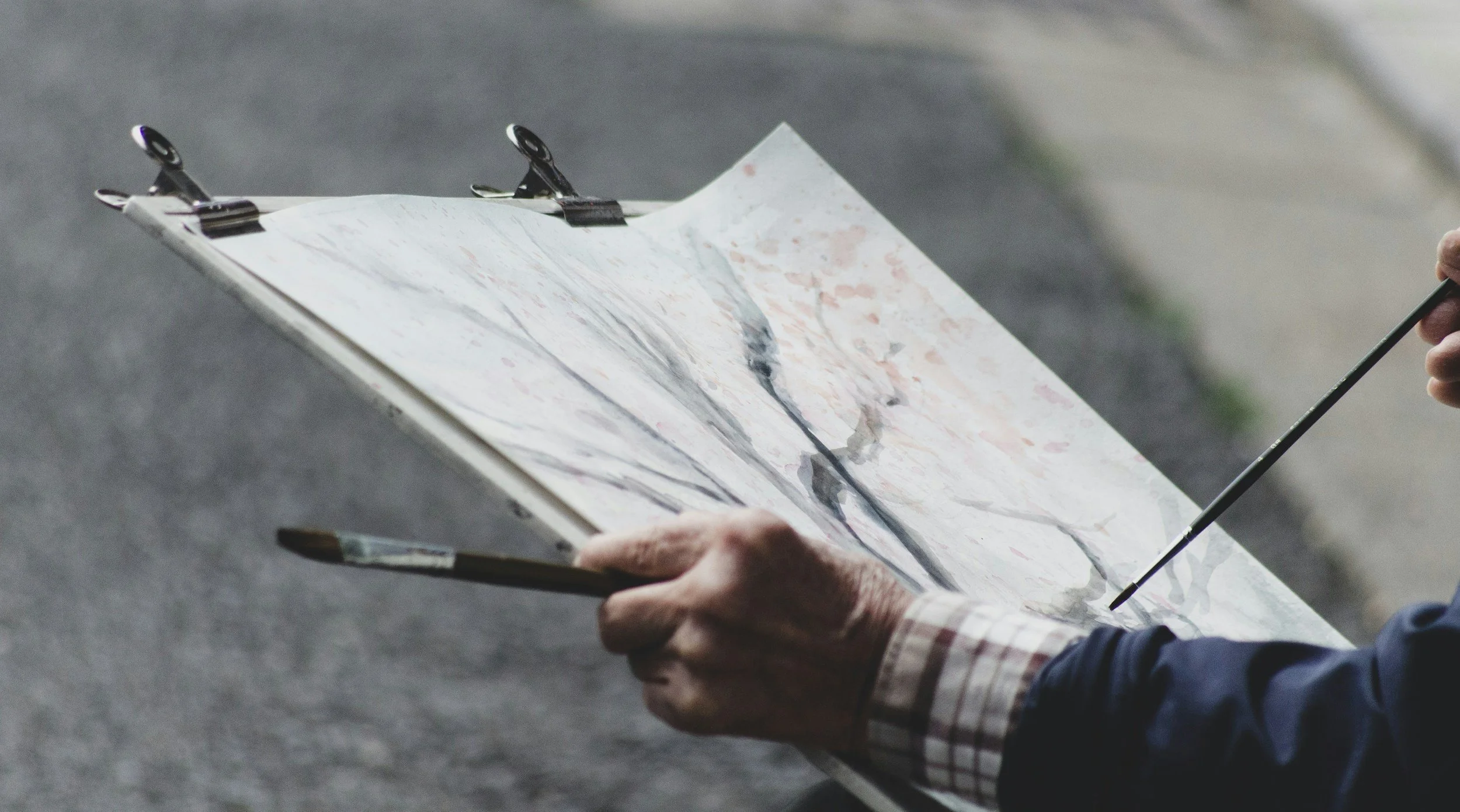 Person painting an abstract watercolor landscape with a thin brush on a sketchboard outdoors.
