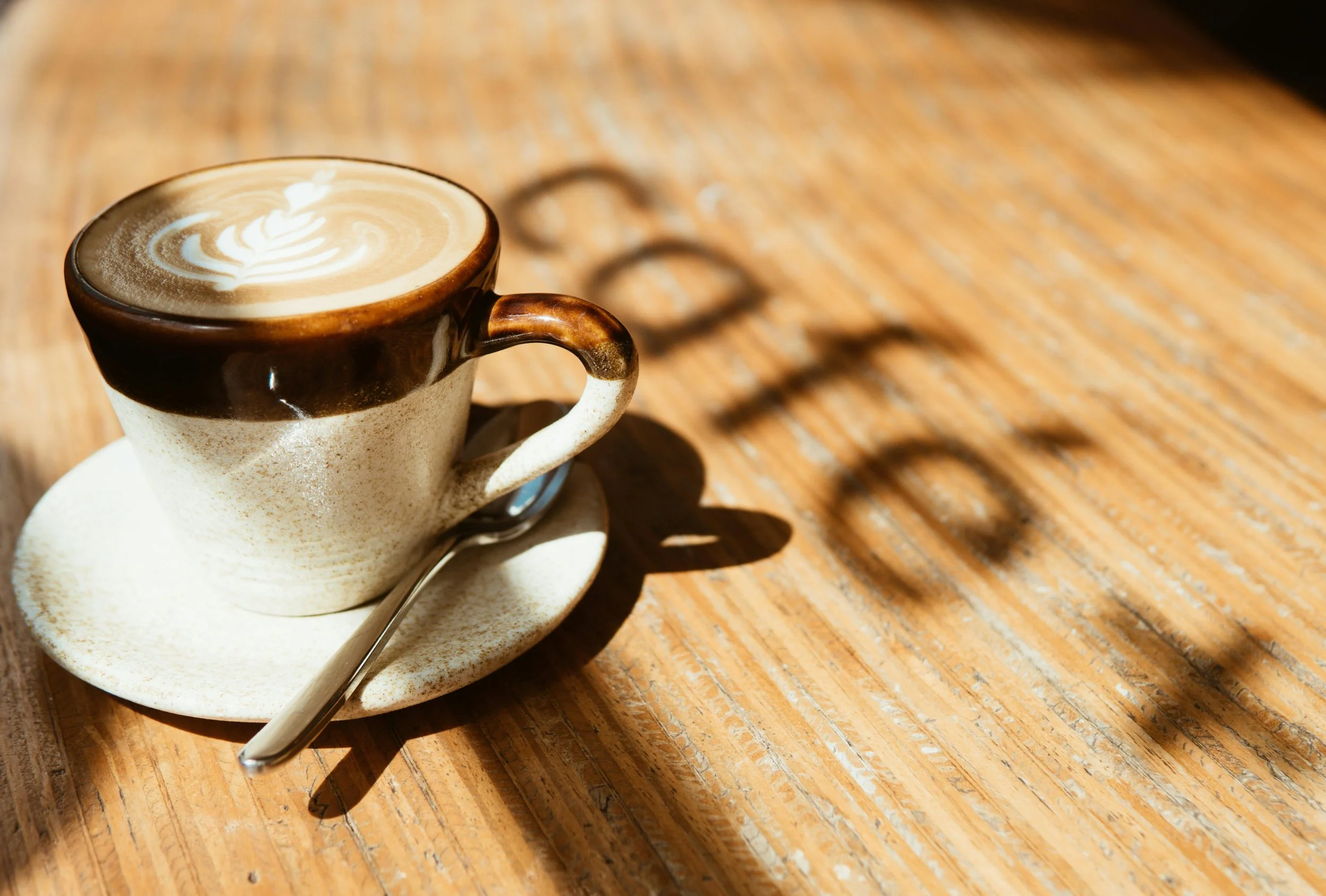 A ceramic cup of coffee with latte art on a wooden table, casting shadows.