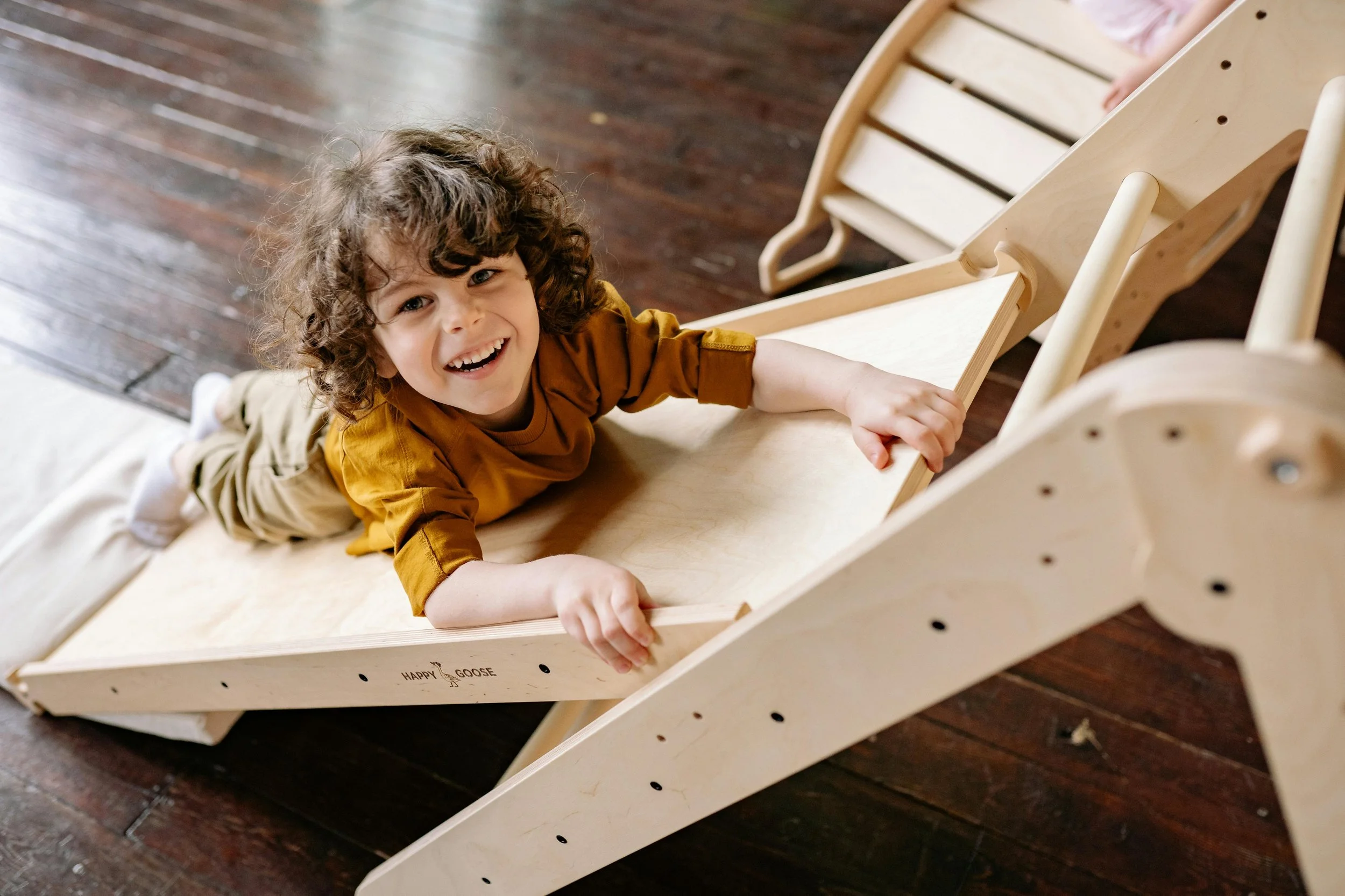 A young child with curly hair and a brown shirt is smiling and climbing an unassembled wooden slide on a wooden floor.