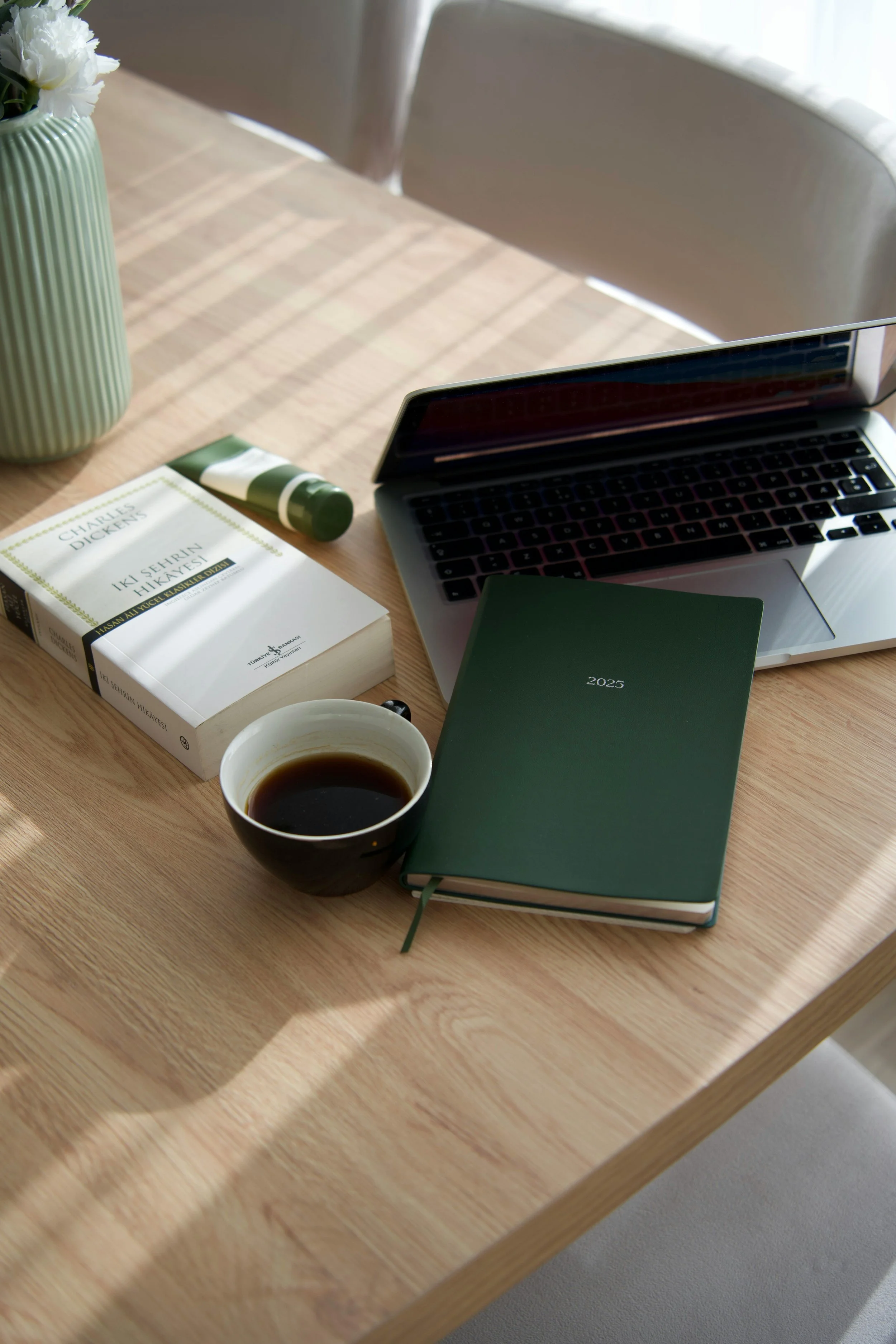 A wooden desk with a black coffee cup, a green 2023 planner, an open silver laptop, a white book titled 'Iki Sehirli Hikayesi', a green and white tube, a green and white vase with white flowers, and a green journal.