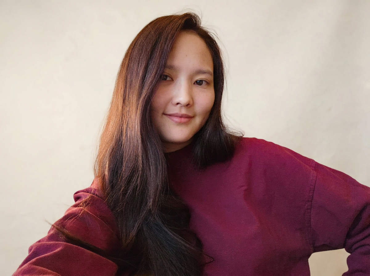 A young woman with long, dark hair and light skin taking a selfie in a maroon top with a neutral background.