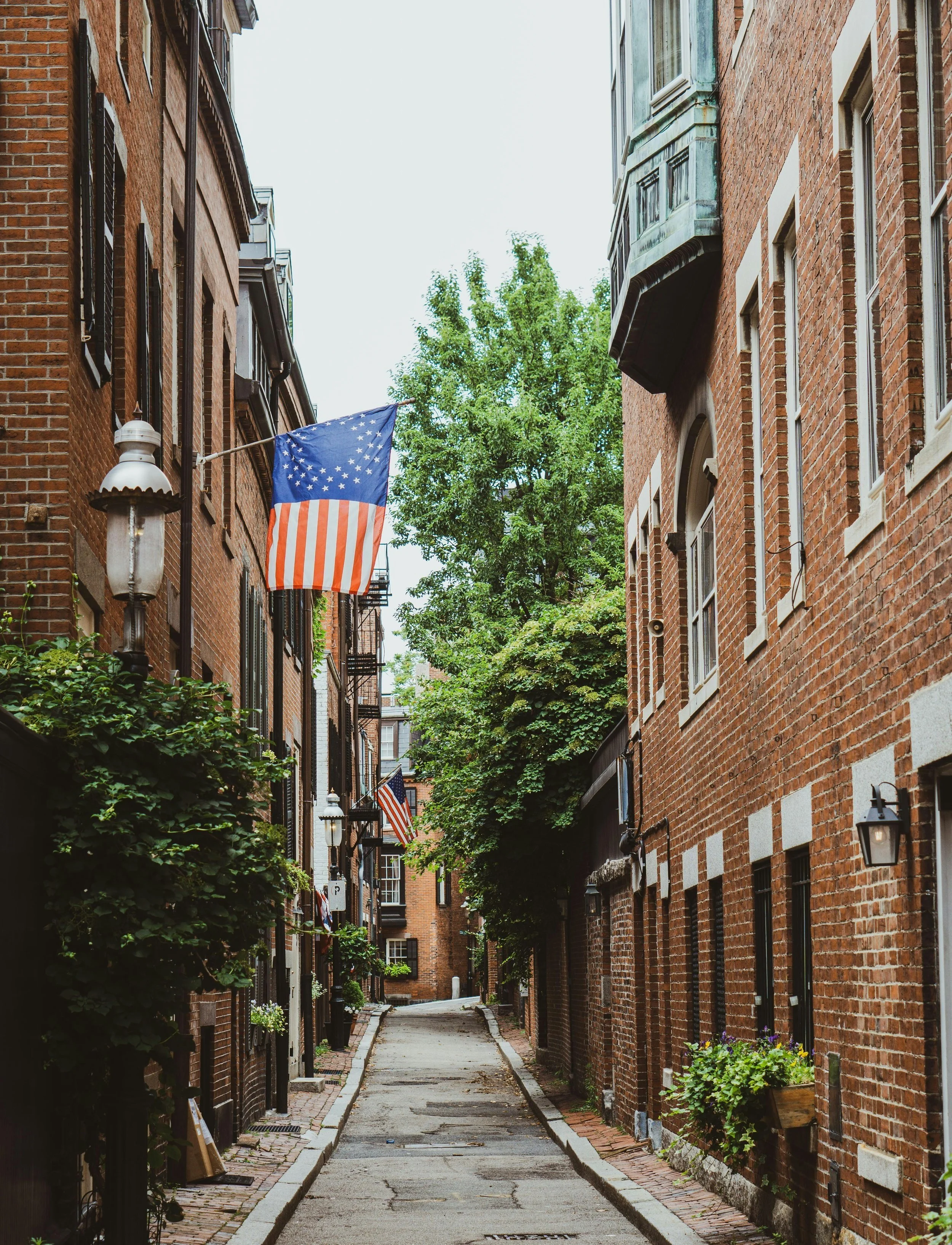 A narrow alleyway between brick buildings decorated with American flags, greenery, and vintage street lamps.