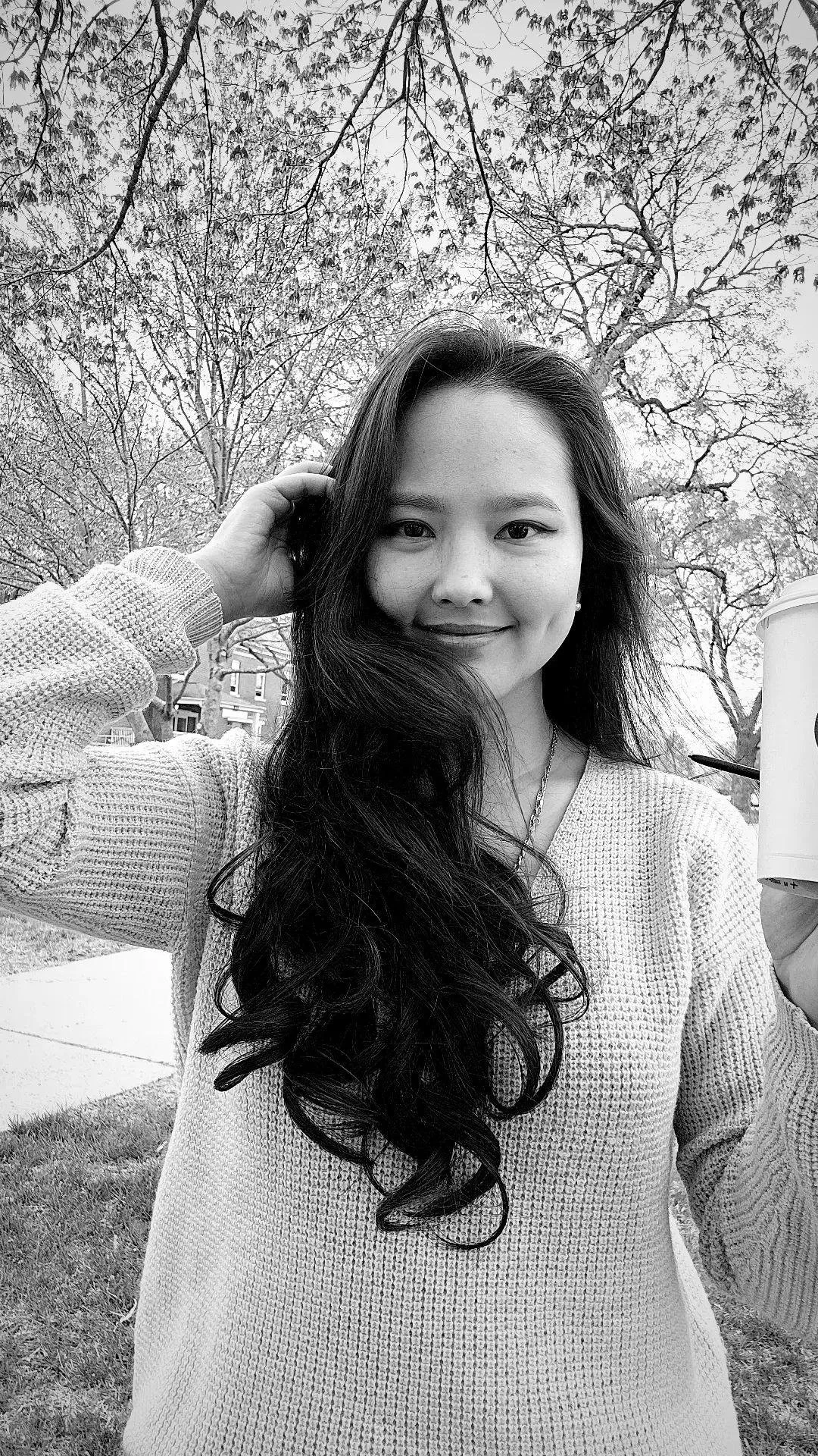 A woman with long dark wavy hair smiling at the camera outdoors in a park, holding a paper cup in her right hand, with leafless trees and houses in the background, in black and white.