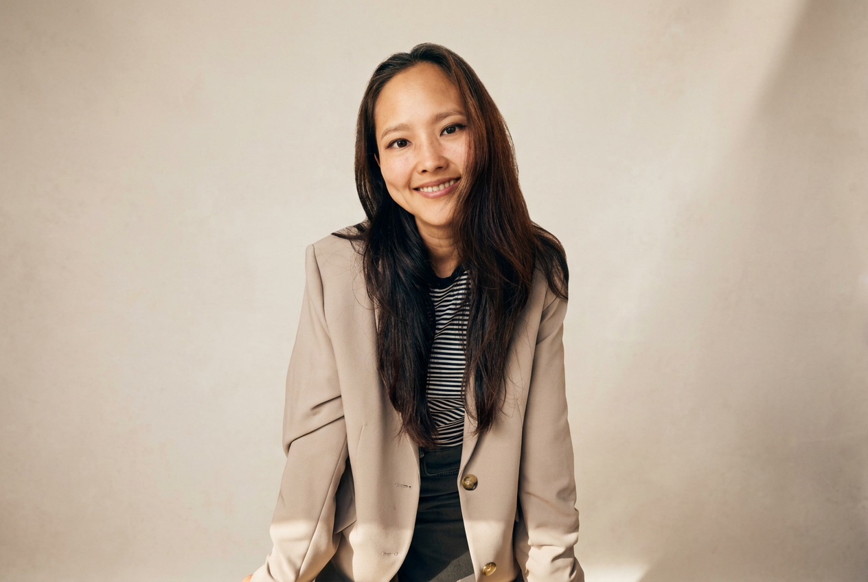 A young woman with long dark hair smiling, wearing a beige blazer over a striped shirt, standing against a plain light-colored background.