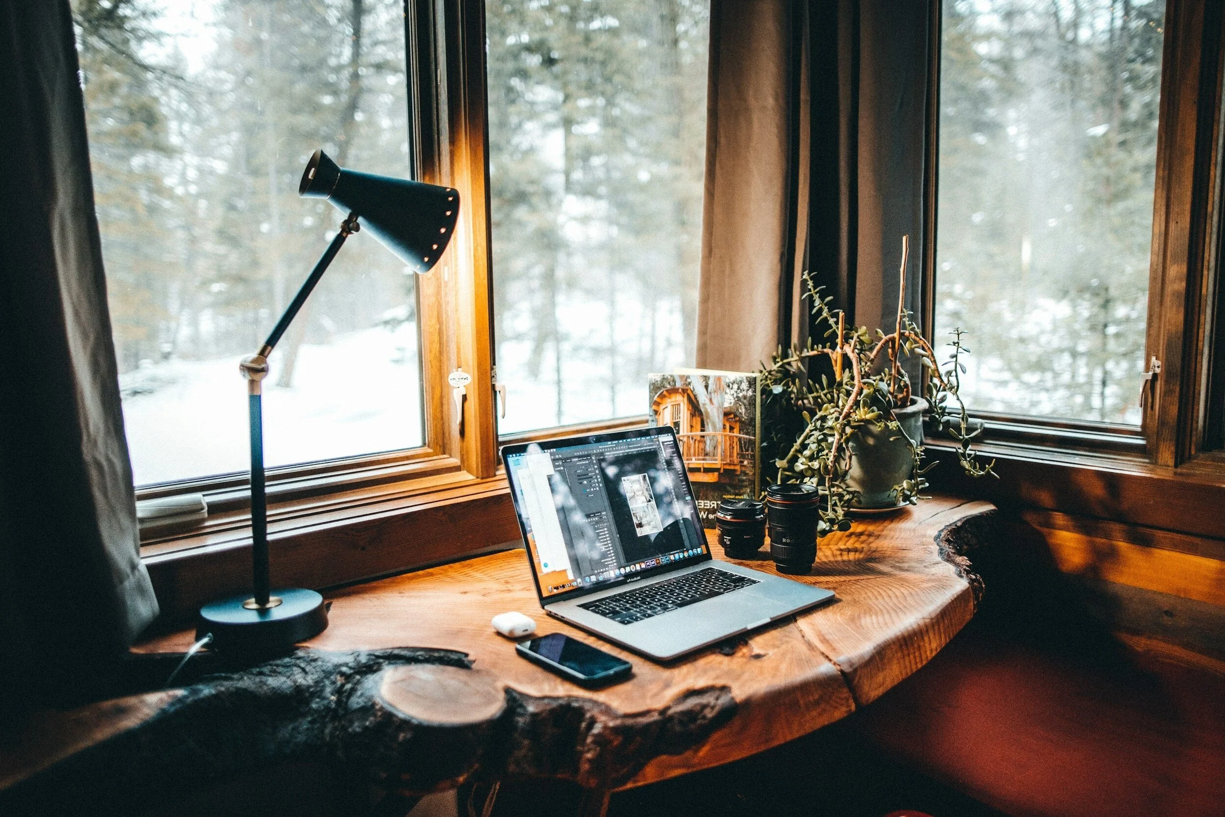 A cozy wooden desk with a laptop displaying photo editing software, a smartphone, a camera lens, a potted plant, and a desk lamp near a large window showing a snowy forest outside.