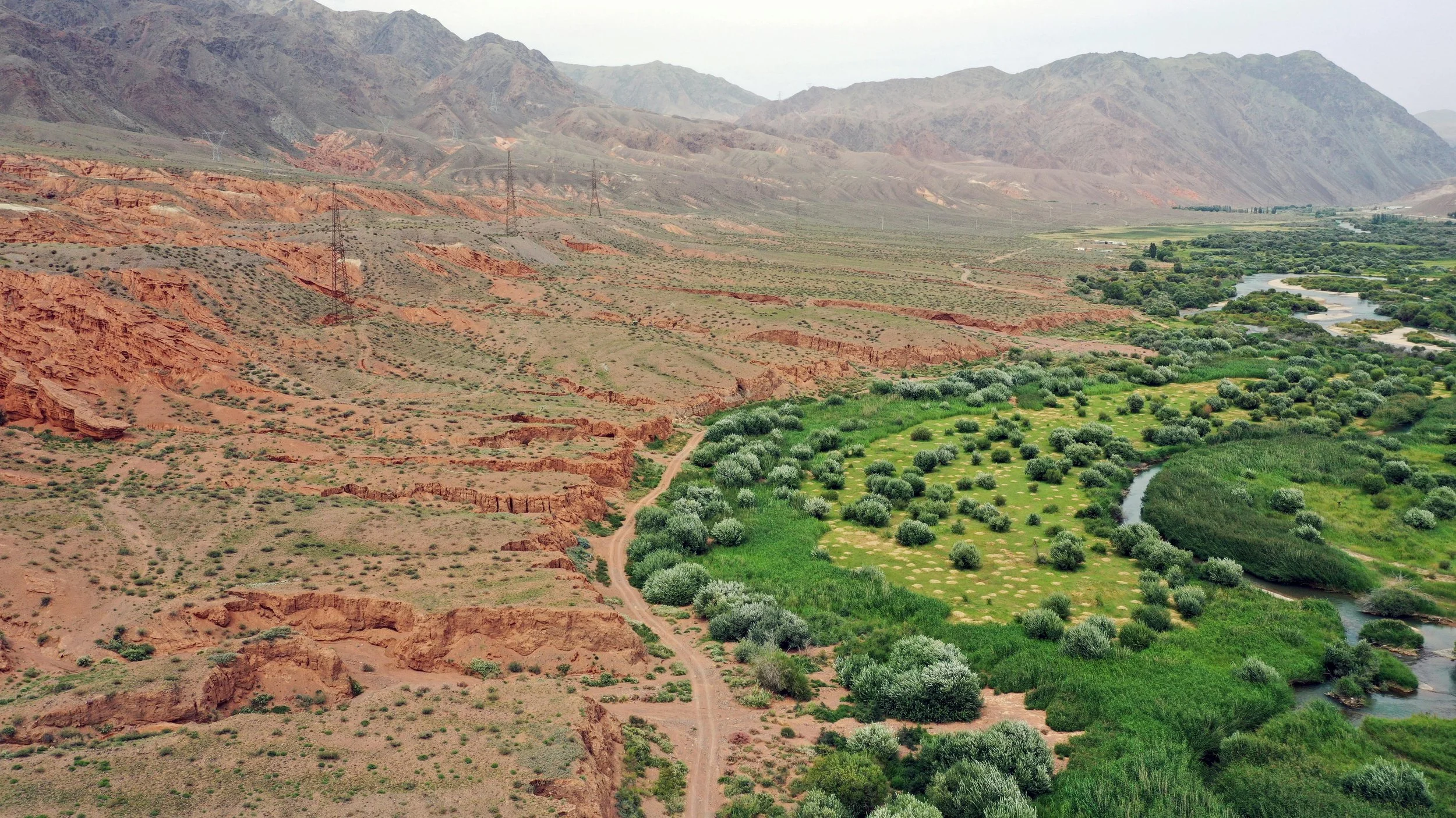 A landscape showing a valley with green vegetation and a winding river, flanked by arid mountains with red and brown hues, and power lines running across the terrain.