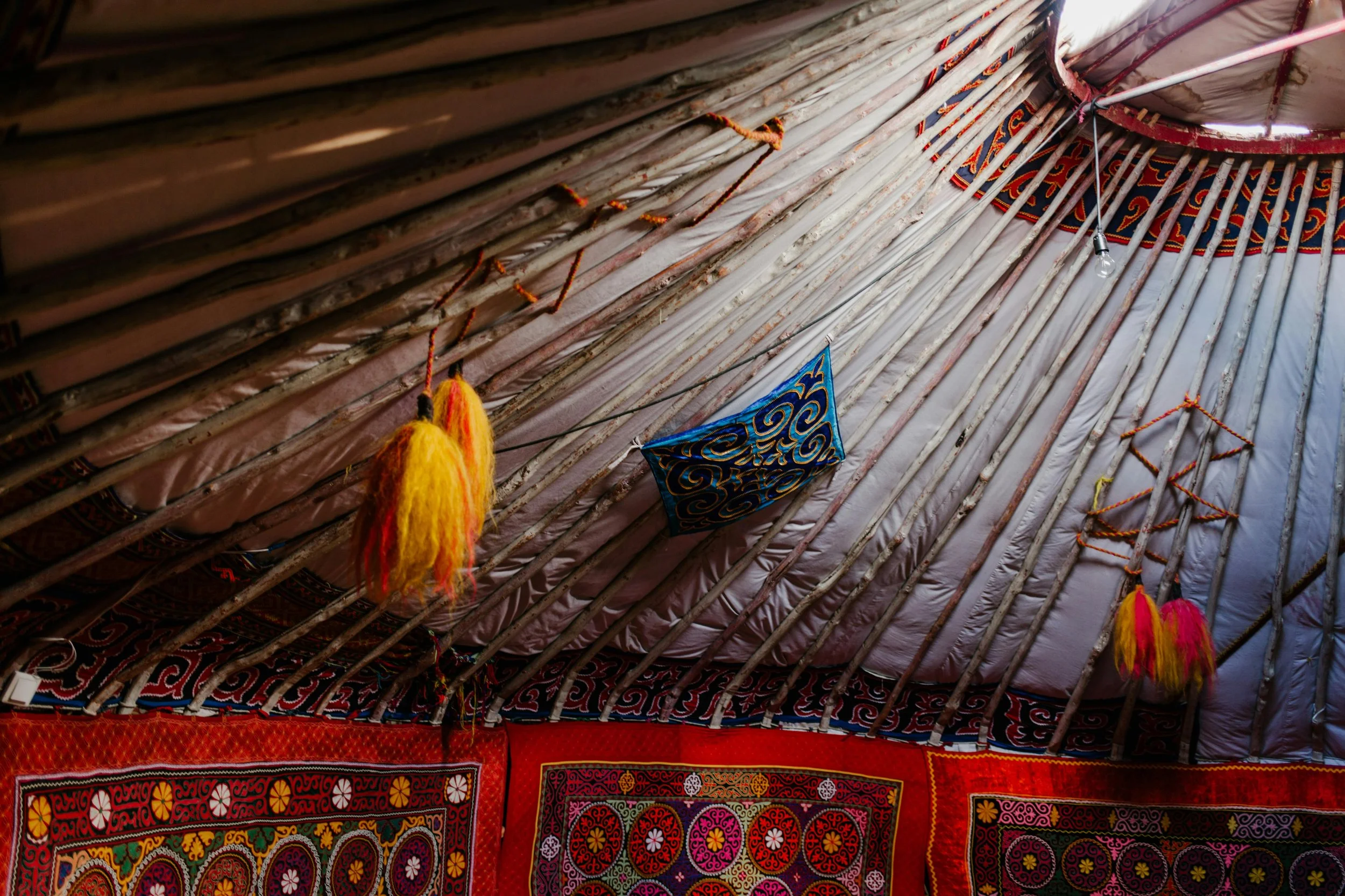 Interior of a traditional yurt with decorated fabric walls and a conical, wooden lattice ceiling, adorned with colorful textiles and hanging decorations.
