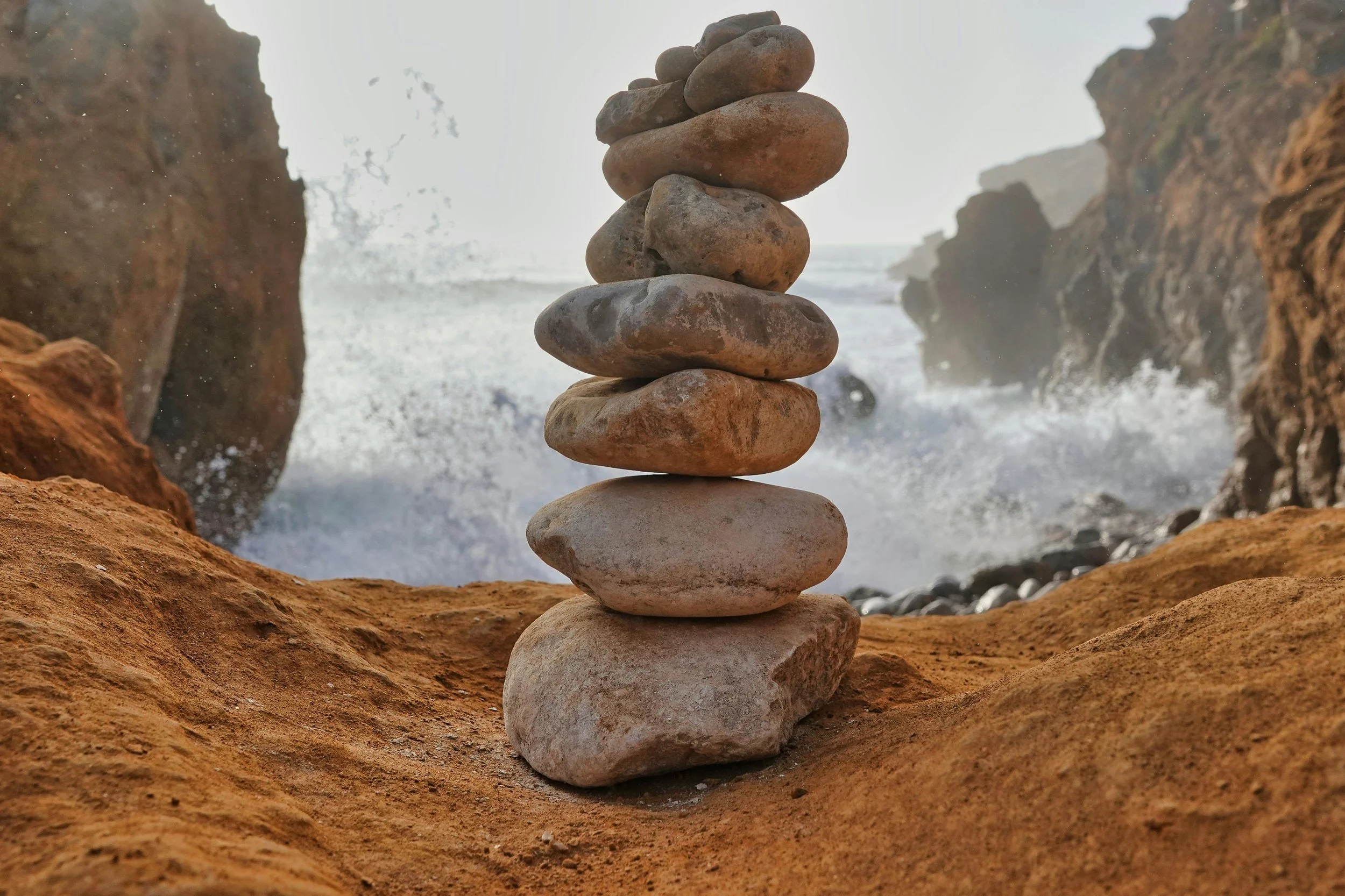 A stack of seven smooth, rounded stones arranged in a balanced tower on a rocky coastal surface with cliffs in the background and ocean waves crashing behind.
