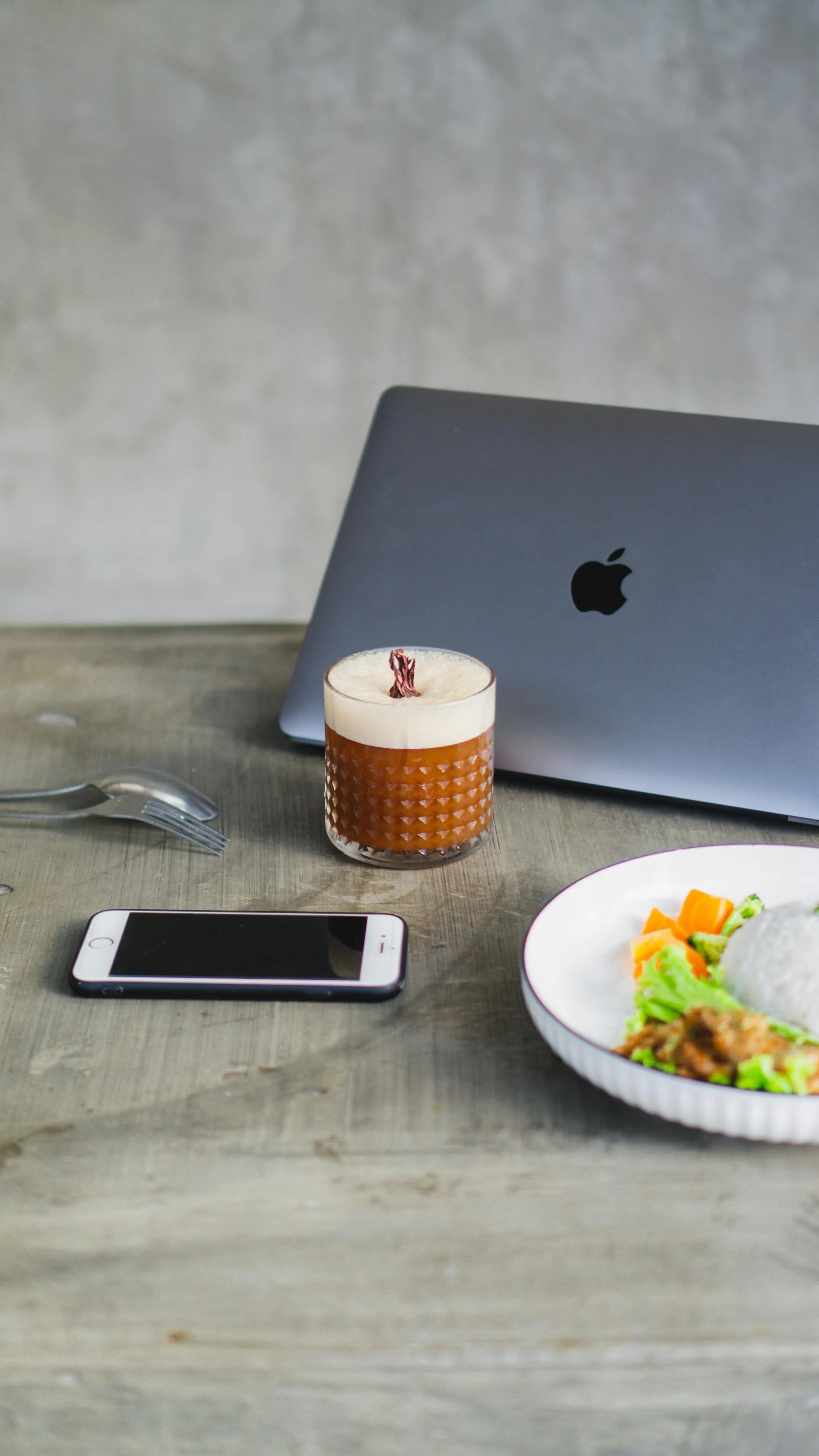 Table with a smartphone, fork, a glass of iced coffee topped with whipped cream, a plate of rice and vegetables, and a closed laptop.