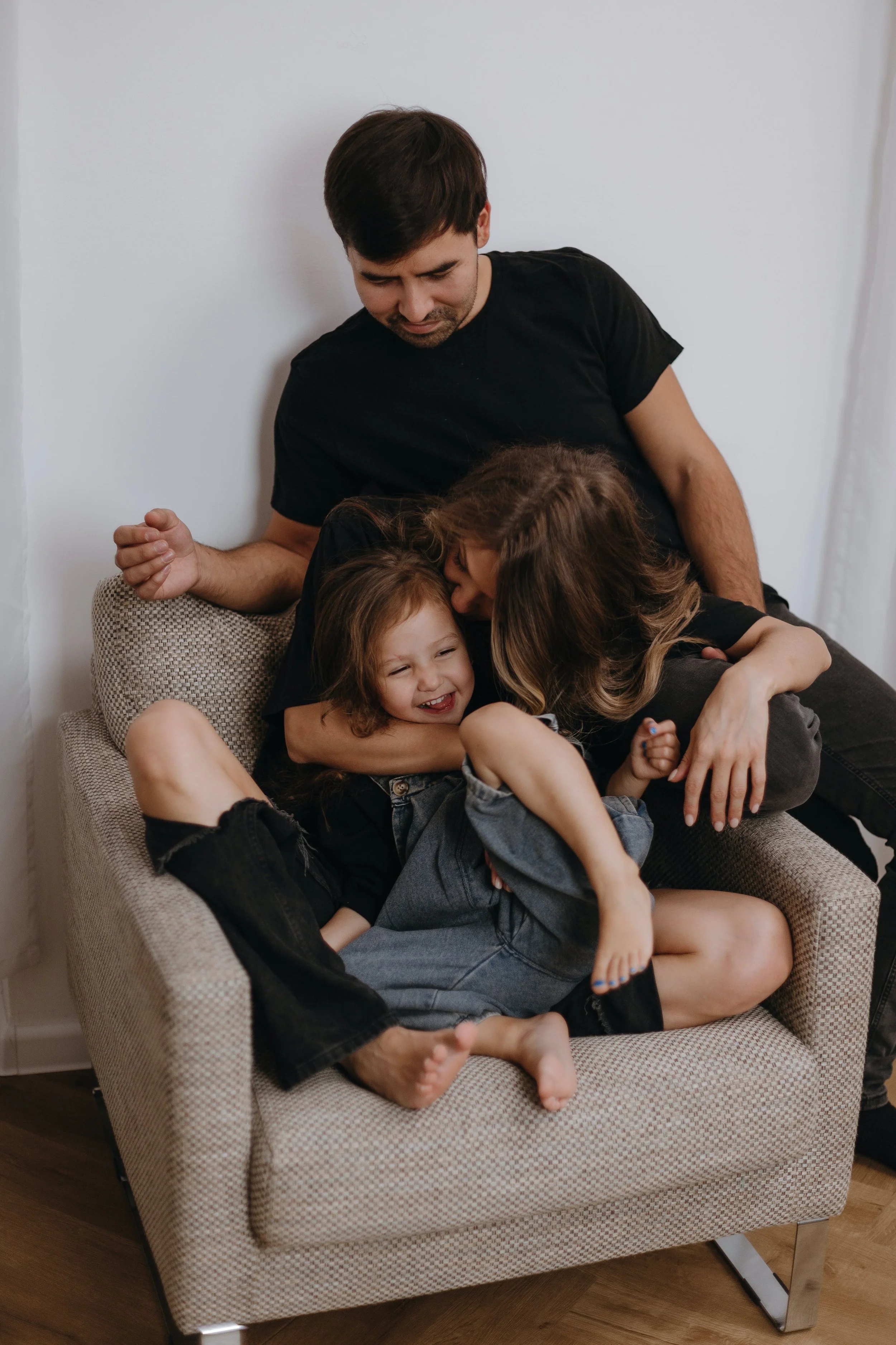 A man and three children cuddling and playing on a beige couch in a living room with white walls.
