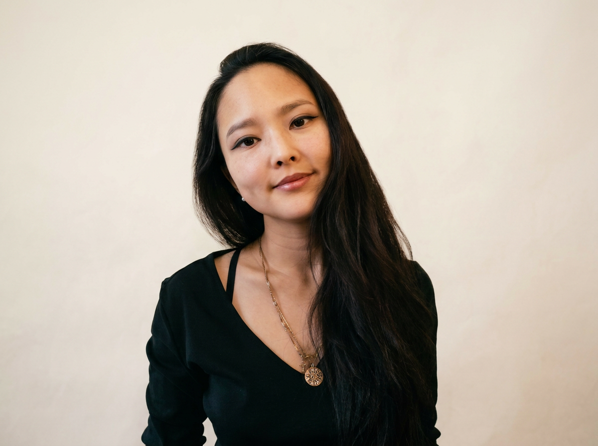 A young Asian woman with long black hair, wearing a black top, gold necklace, and earrings, posing against a plain light-colored wall.