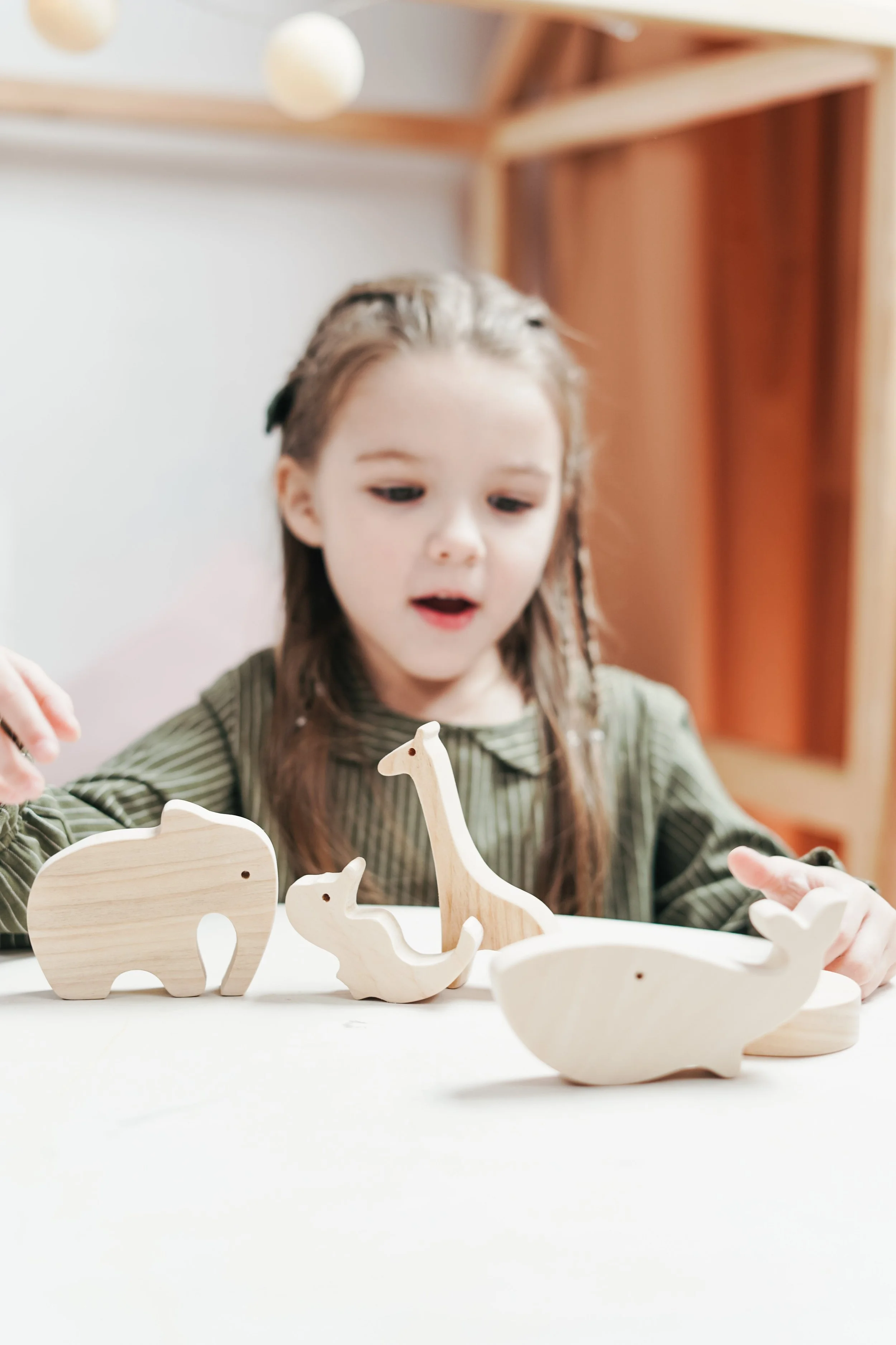 A young girl with long brown hair, wearing a green striped shirt, looking at wooden animal figurines on a table, including an elephant, giraffe, and whale, in a room with beige walls and wooden shelves.
