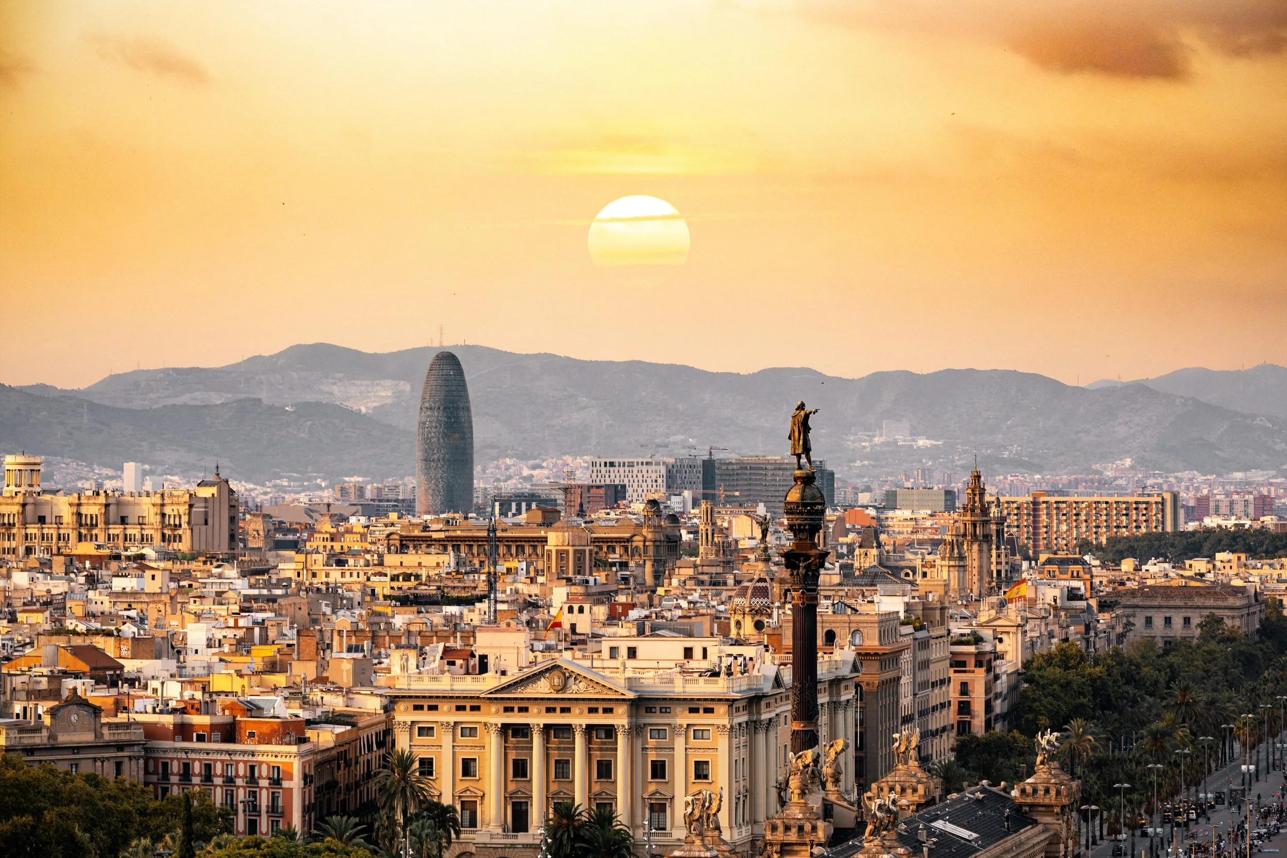 Cityscape of Barcelona at sunset, featuring the Sagrada Família cathedral, the Christopher Columbus monument, and the Torre Agbar building with mountains in the background.