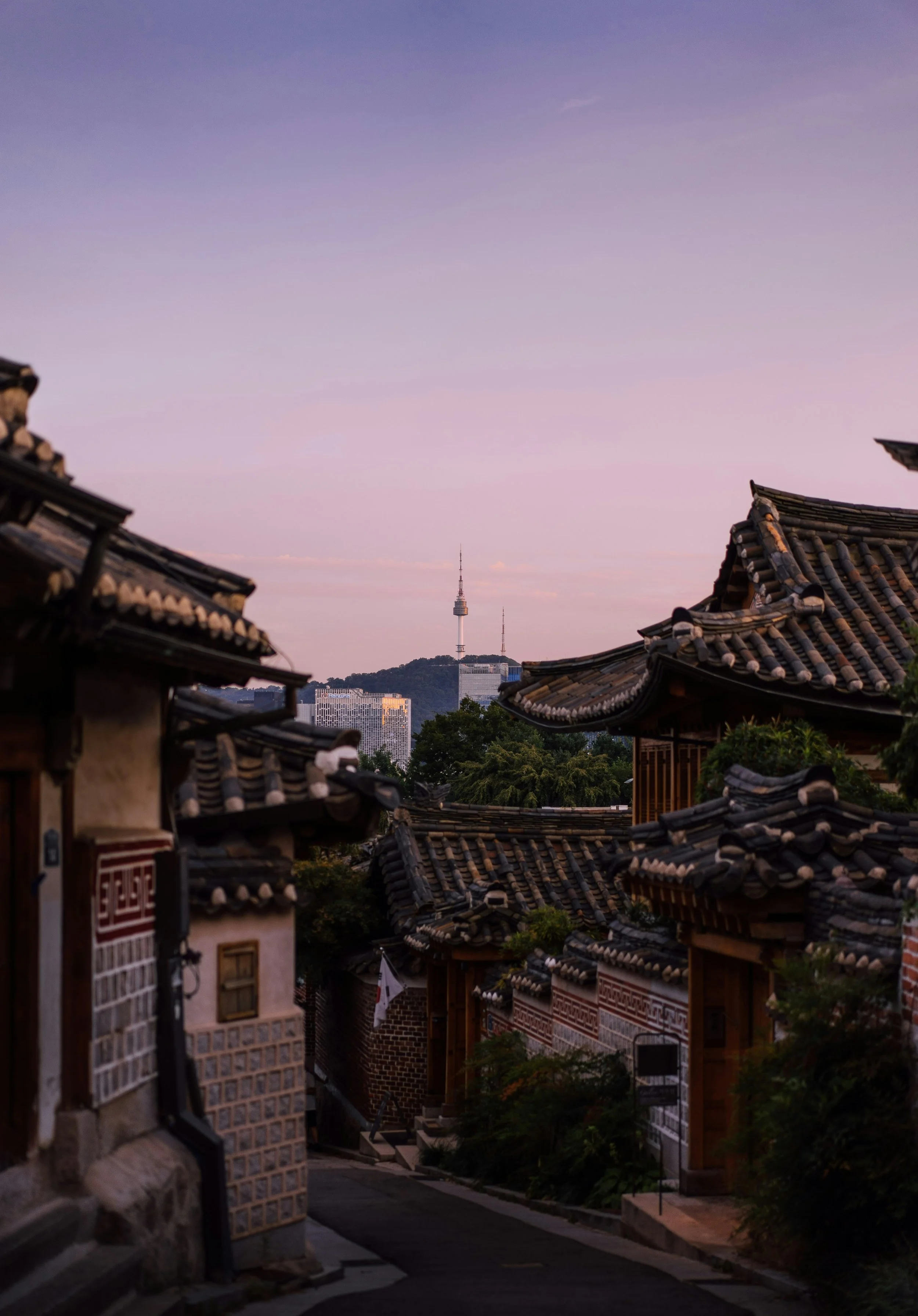 Traditional Korean hanok rooftops with modern Seoul city skyline and Namsan Seoul Tower in the background during early evening.