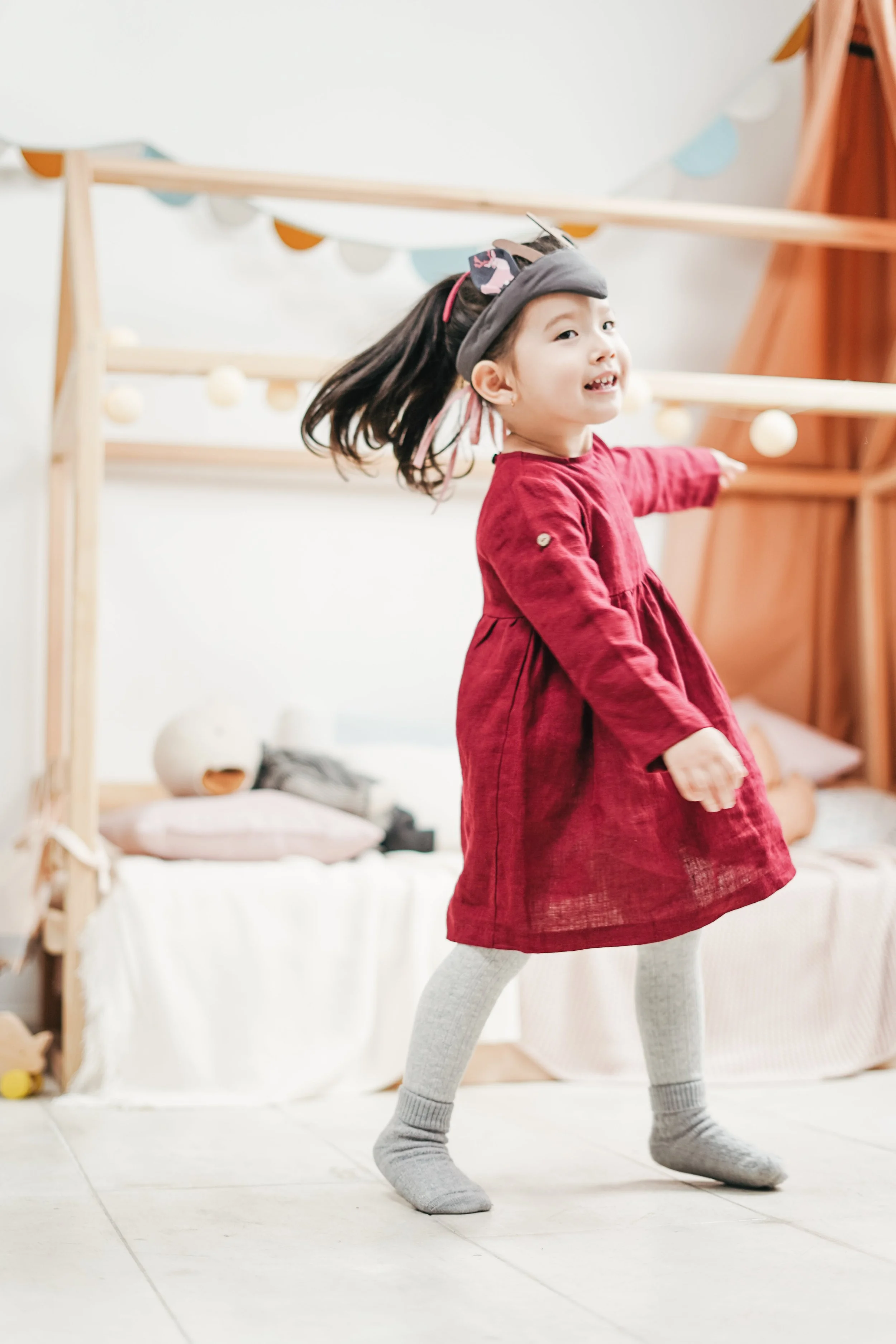 Young girl in a red dress and gray socks playing indoors, balancing on her toes, with a bed and toys in the background.