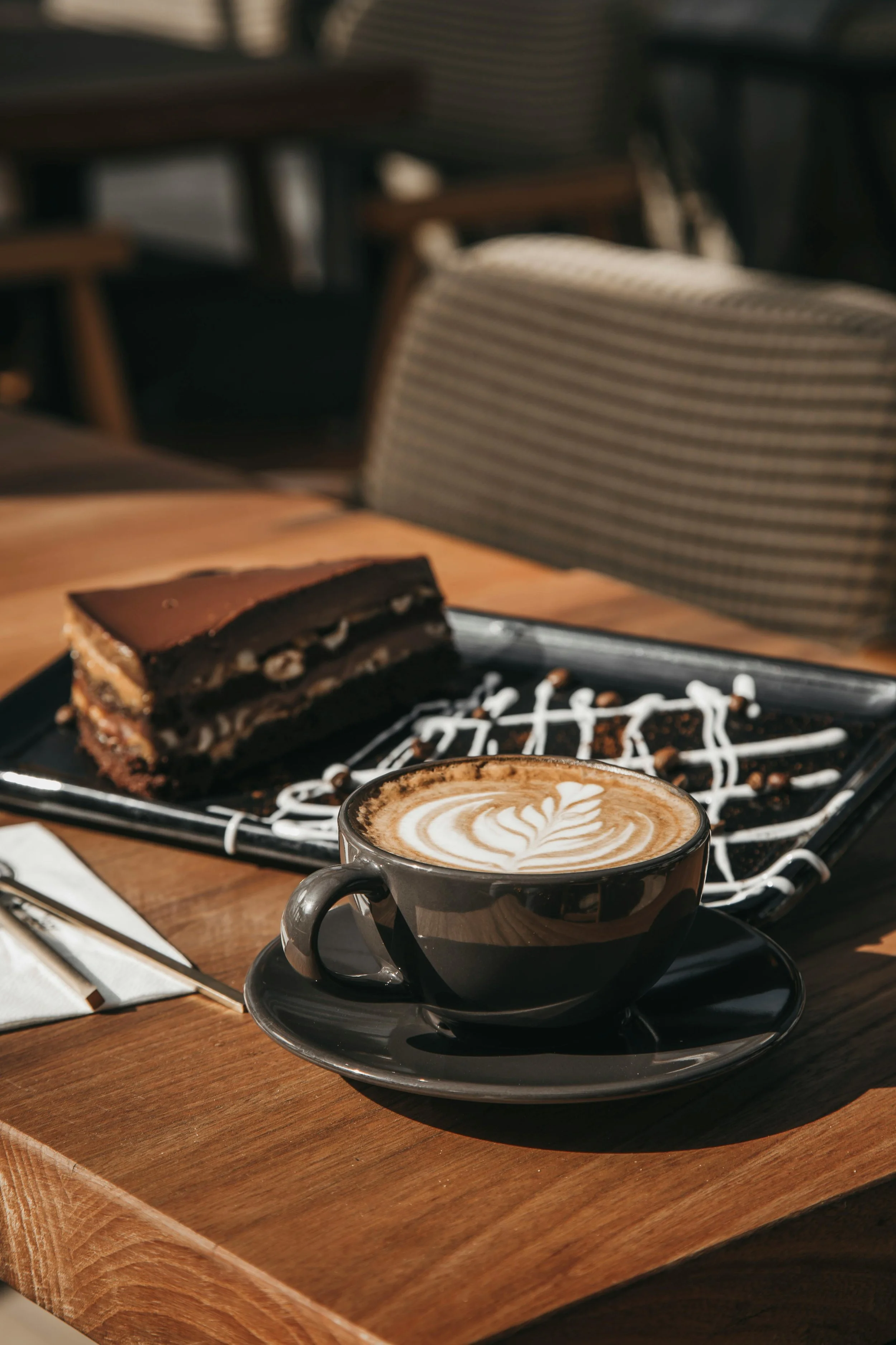 A cup of latte with foam art on a black saucer, and a slice of chocolate cake on a black plate, on a wooden table in a cozy cafe setting.