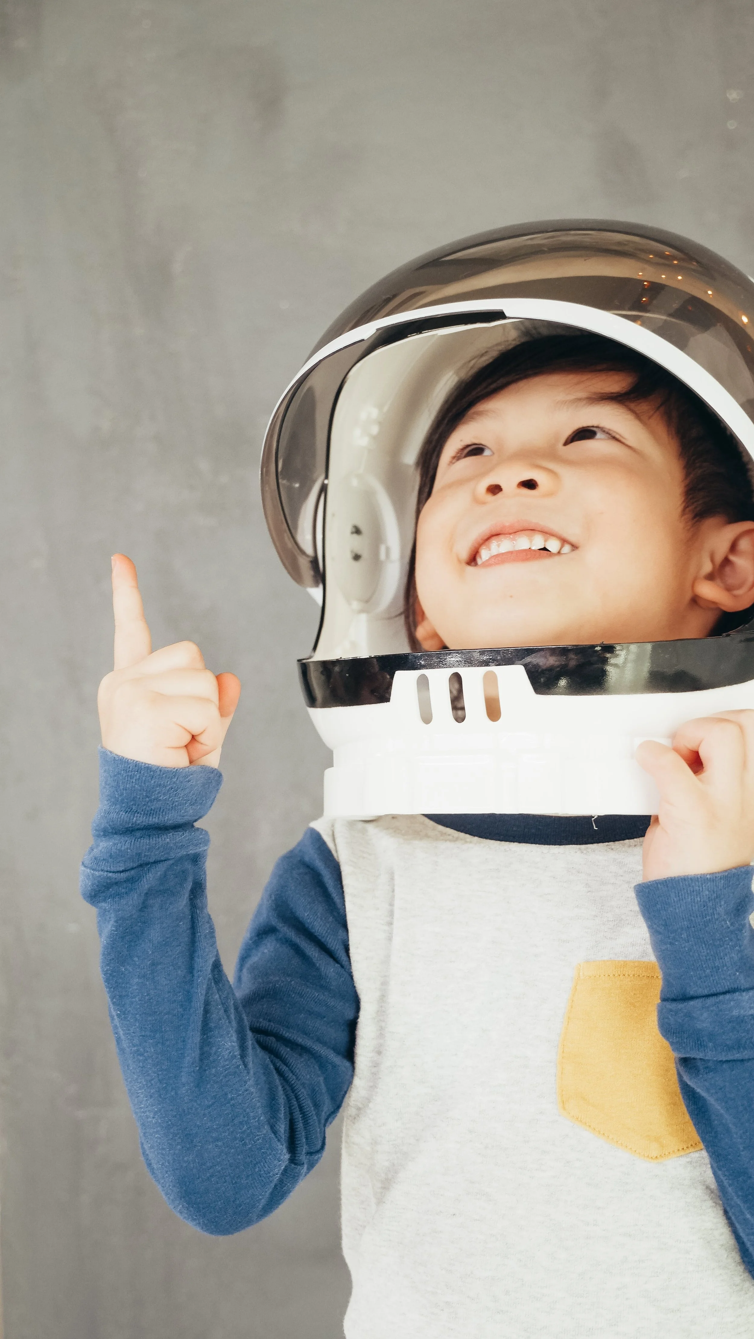 A young boy smiling and pointing upward while wearing an astronaut helmet costume. He is dressed in a long-sleeve shirt with a color block pattern and stands against a neutral gray background.