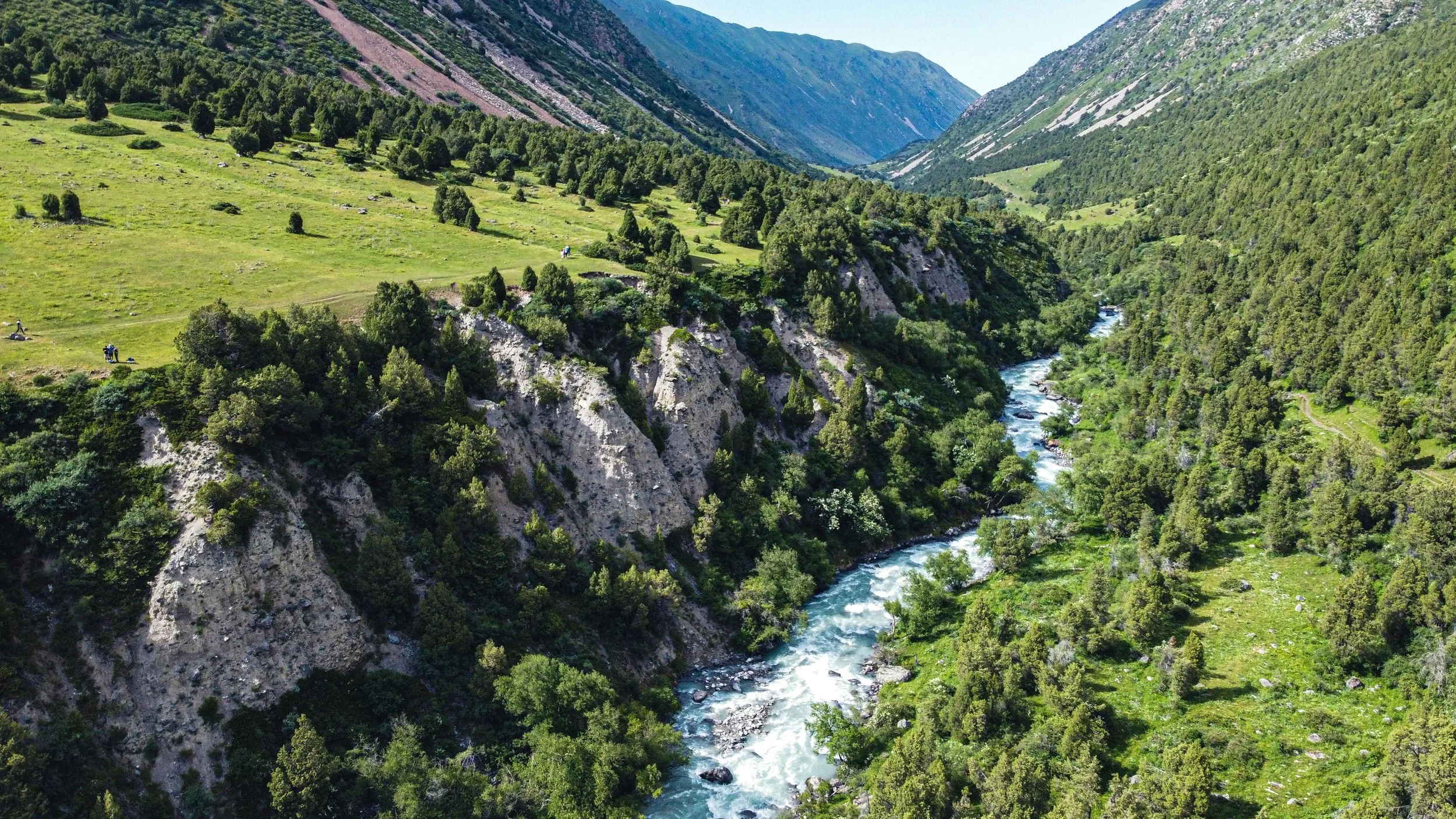 A scenic view of a winding river flowing through a lush green valley surrounded by steep mountains covered in trees and grass.