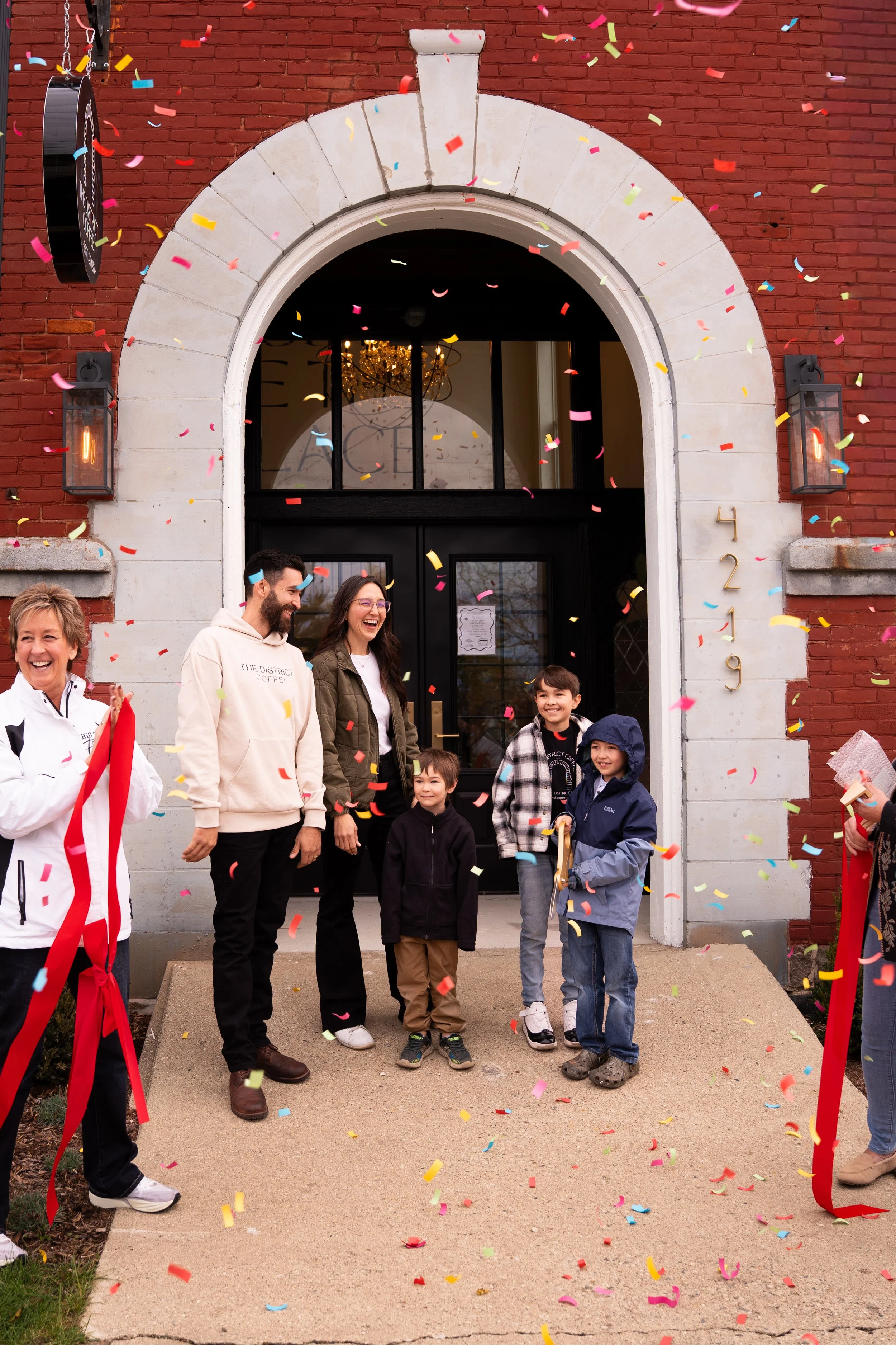 Group of people celebrating a grand opening or special occasion in front of a brick building, with confetti falling and smiling faces.