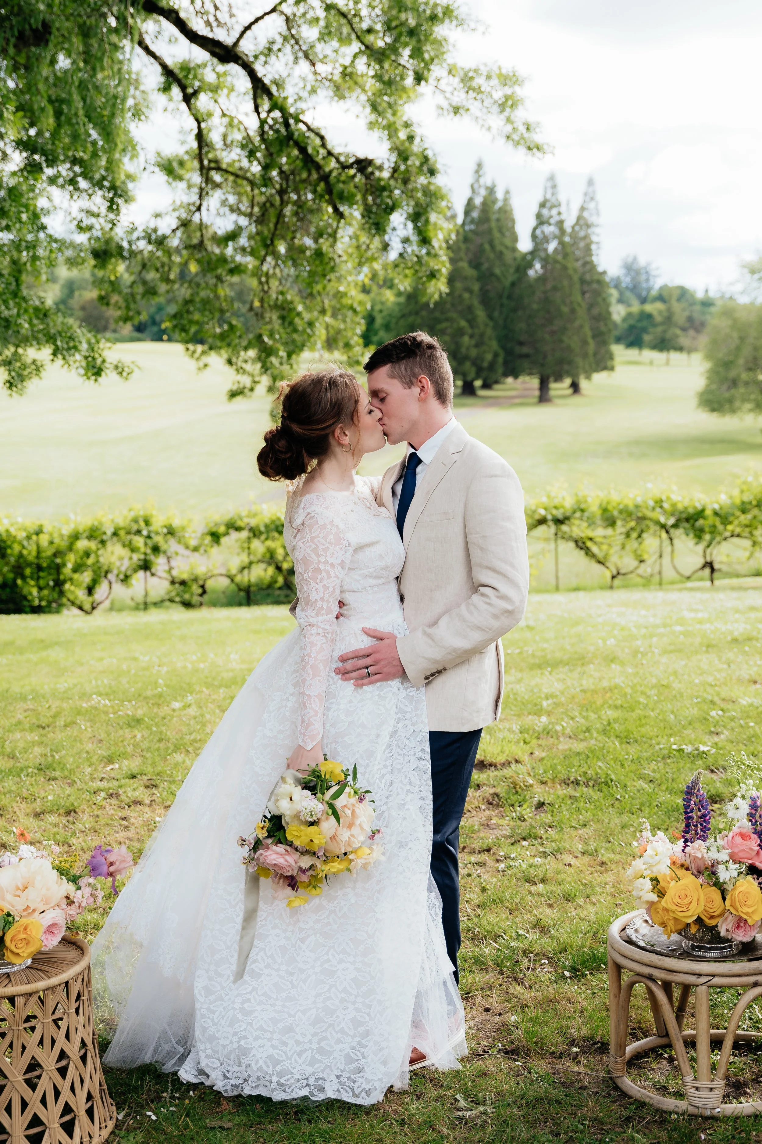 A bride and groom share a kiss outdoors with greenery and trees in the background. The bride holds a bouquet of flowers, and there are flower arrangements on small tables beside them.