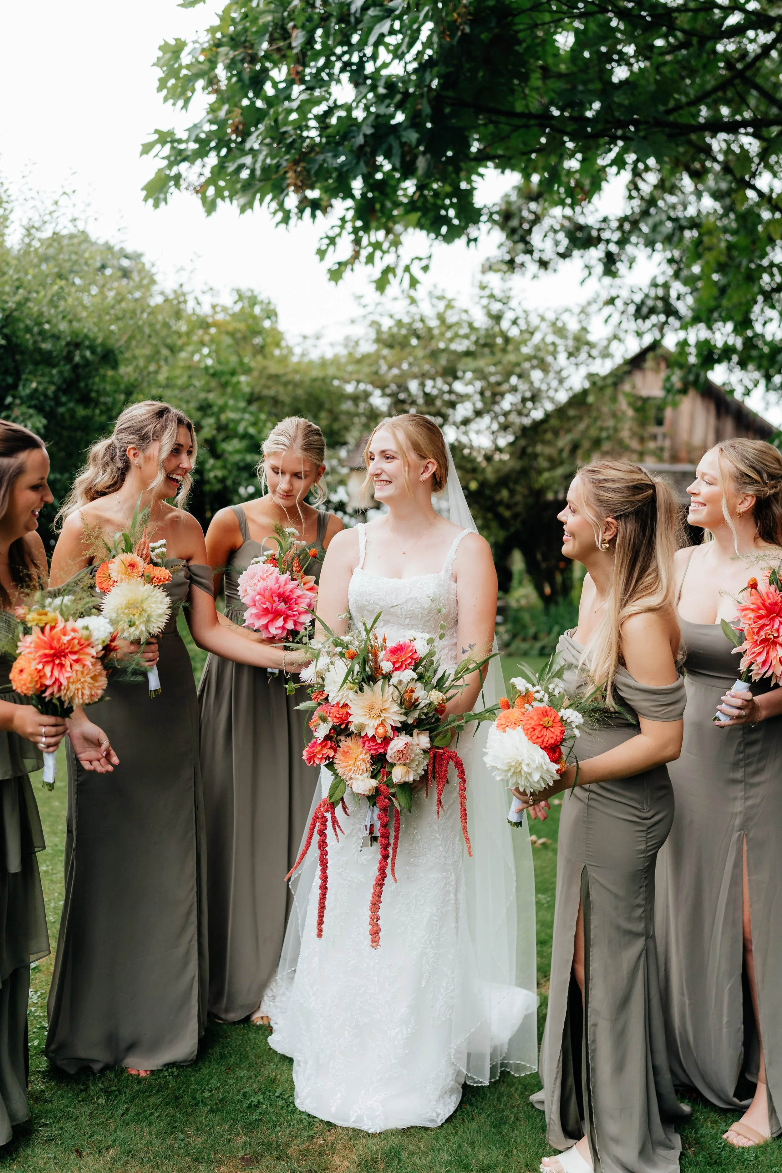 A bride in a white wedding dress and veil holding a large bouquet, surrounded by bridesmaids in sage dresses holding flowers, outdoors with trees in the background.