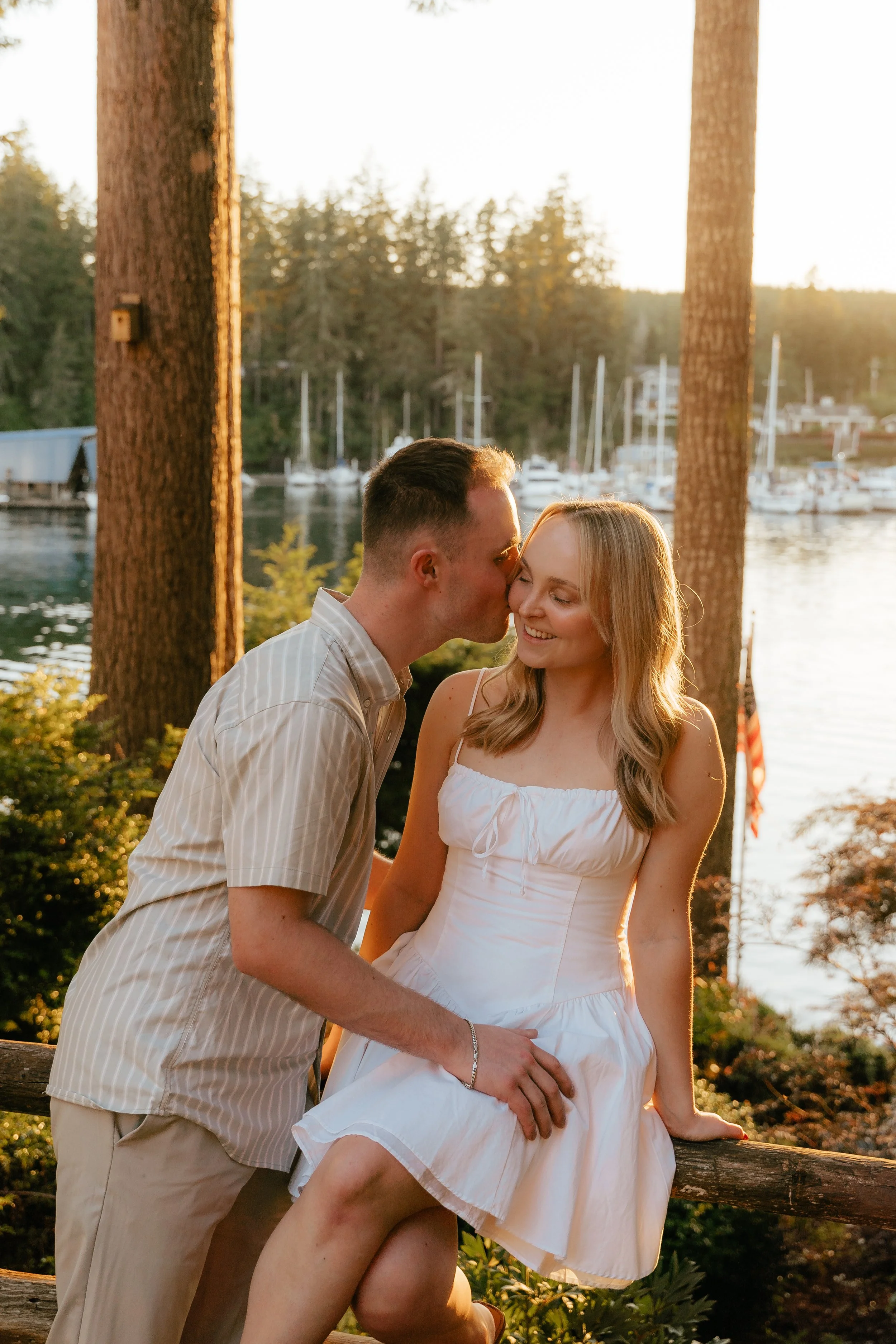 A young couple sitting outdoors near a lake during sunset. The man is kissing the woman on the forehead, and she is smiling with her eyes closed. There are boats and trees in the background.