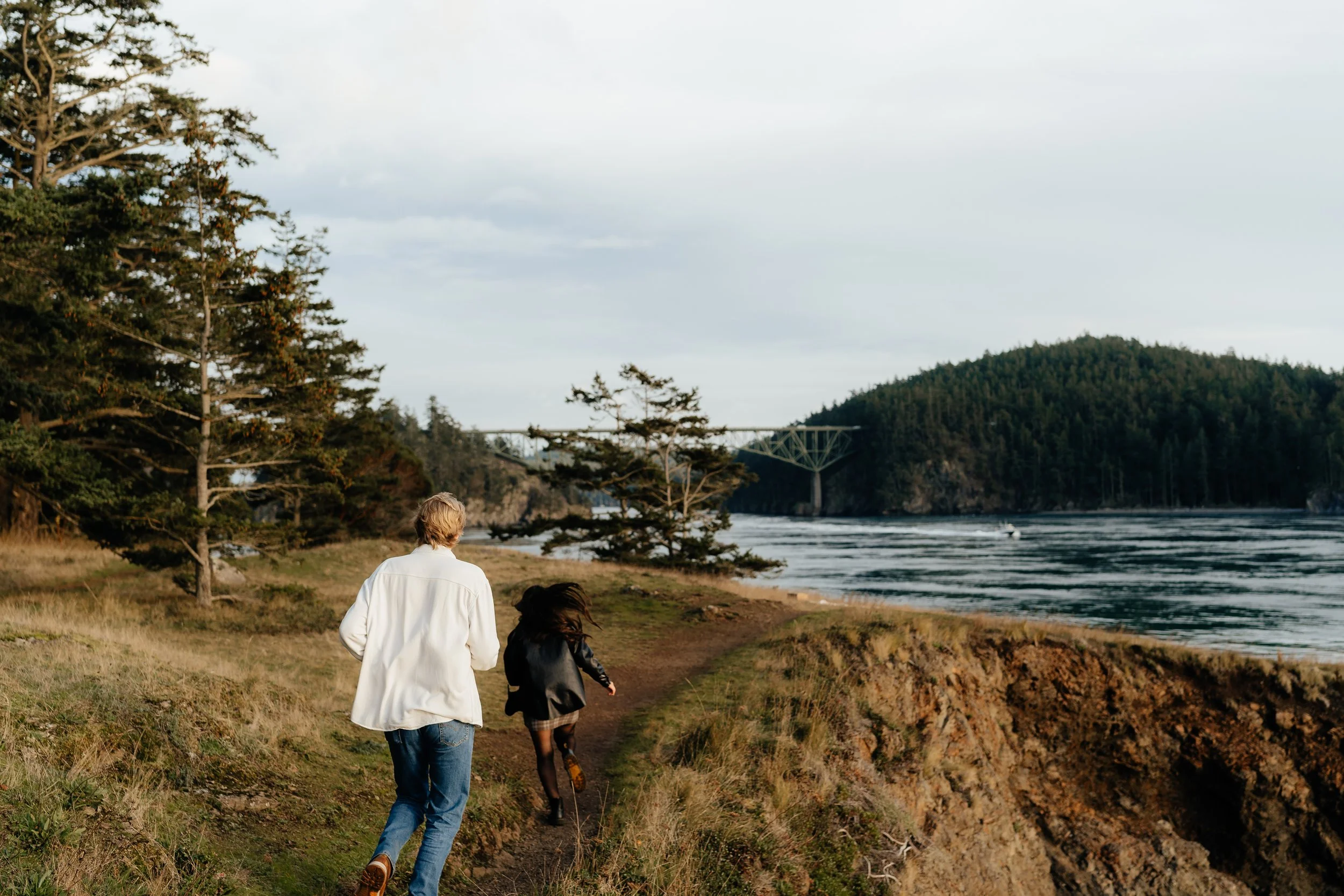 An engaged couple are running along a dirt path beside the ocean, with trees and a bridge in the background.