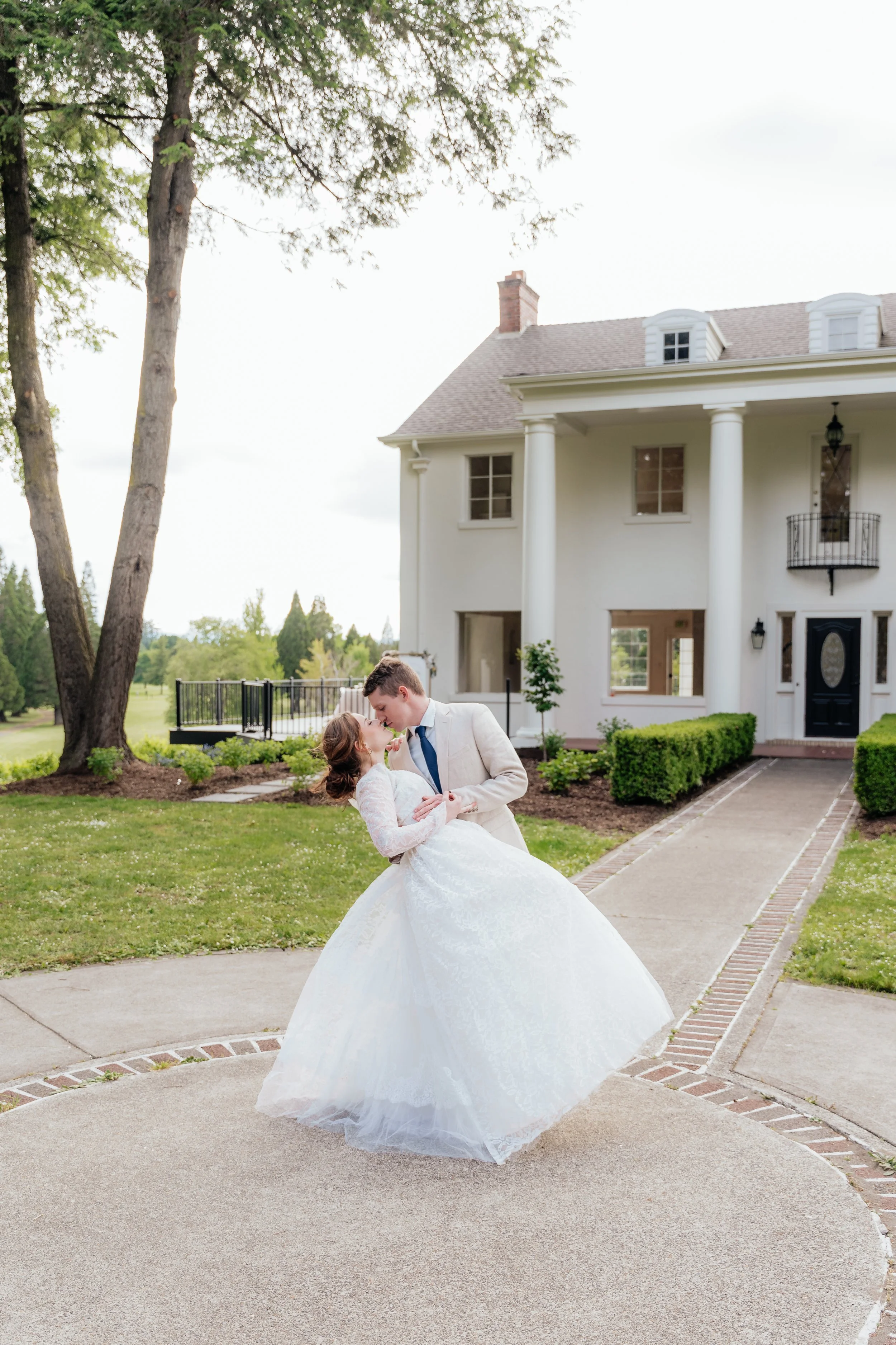 A young man in a beige suit and a young woman in a white wedding gown sharing a dance outside a large white house with tall columns, green bushes, and trees in the background.