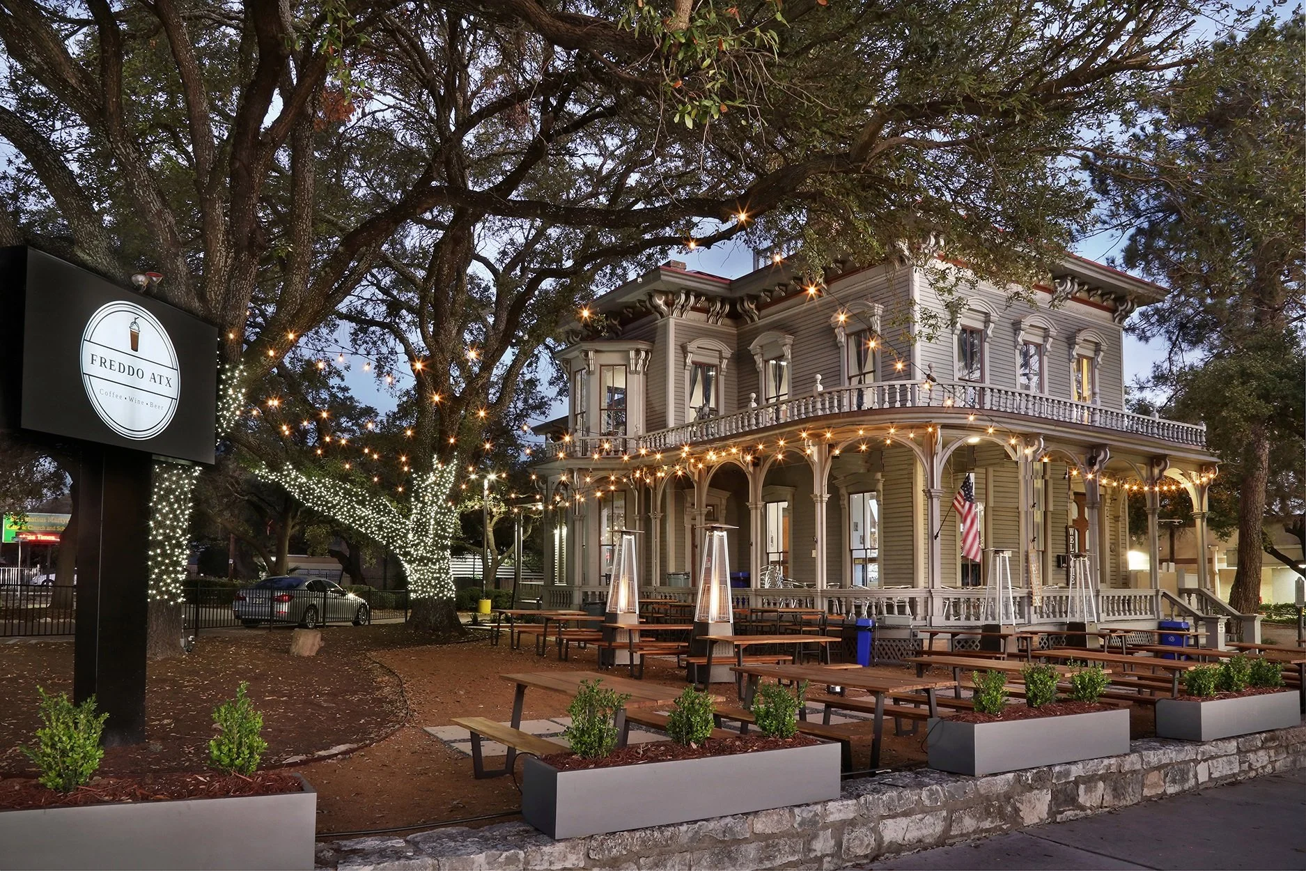 A two-story Victorian-style building with a large porch, string lights, and an American flag, surrounded by trees decorated with string lights, with outdoor seating and a sign that reads "FREDDO ATX".