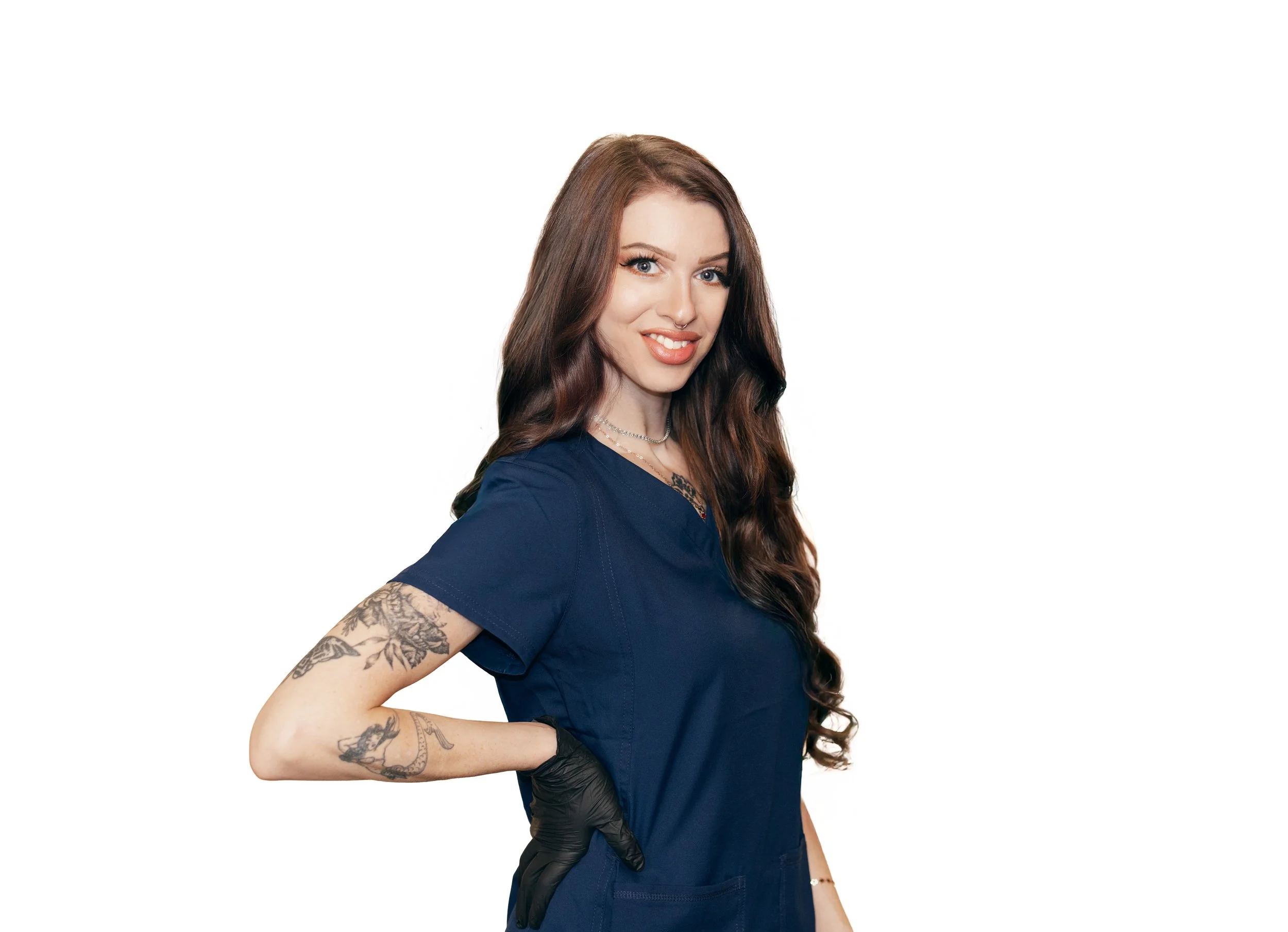 A woman with long wavy brown hair in a blue medical scrub top, smiling, with tattoos on her left arm and wearing a black glove on her right hand, standing against a white background.