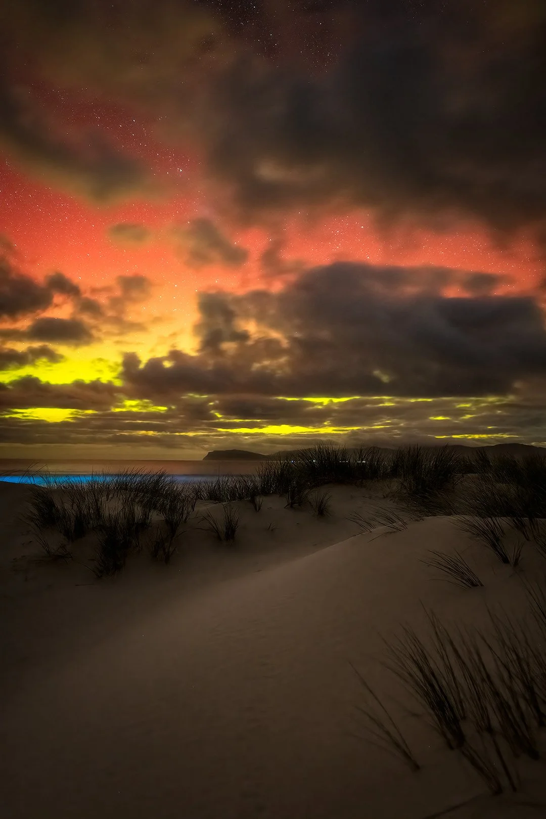 Nighttime scene of sandy dunes with grass, colorful sky with pink, red, yellow, and dark clouds, and stars visible in the sky.