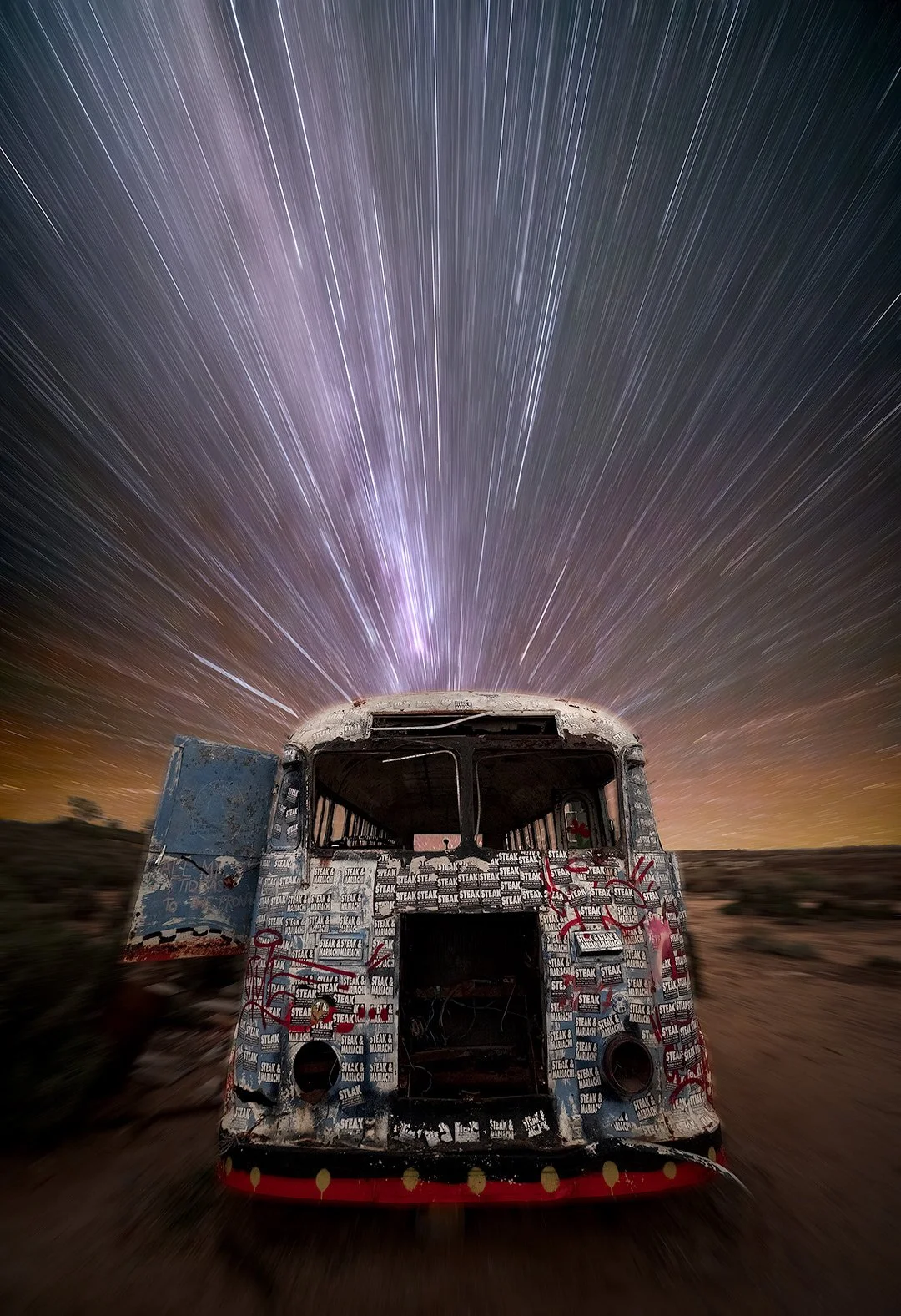 Old abandoned bus with graffiti in a desert landscape under a starry night sky with star trails.